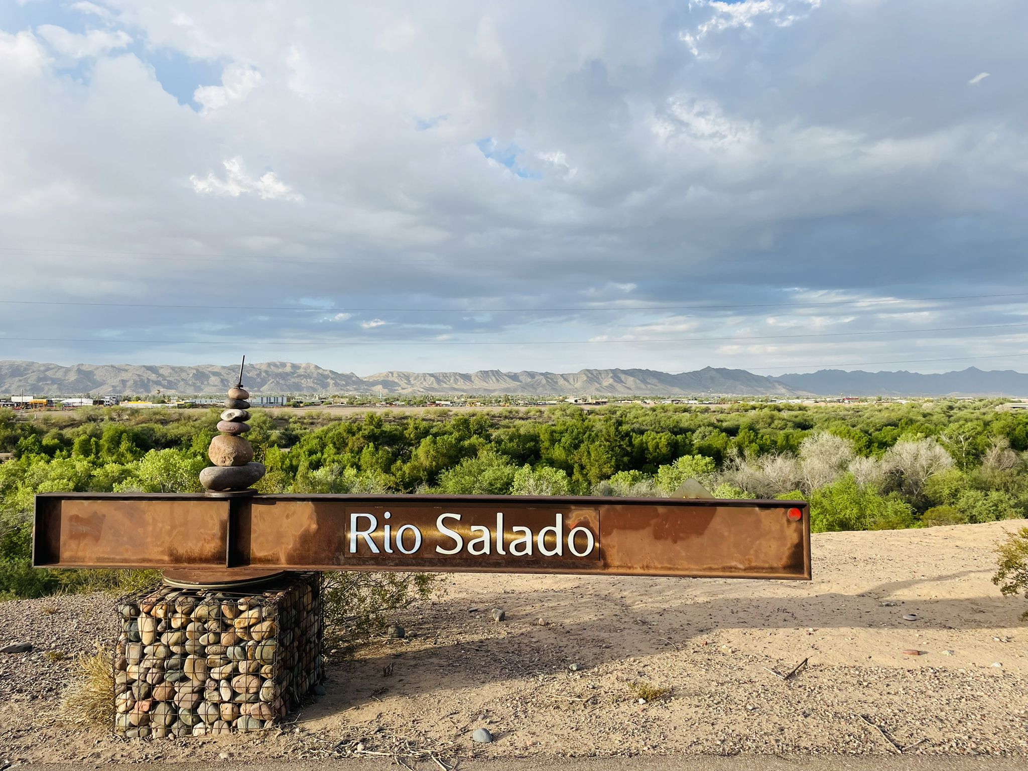 Metal sign with stacked rocks overlooks a green landscape under cloudy skies. - Rio Salado Pathway