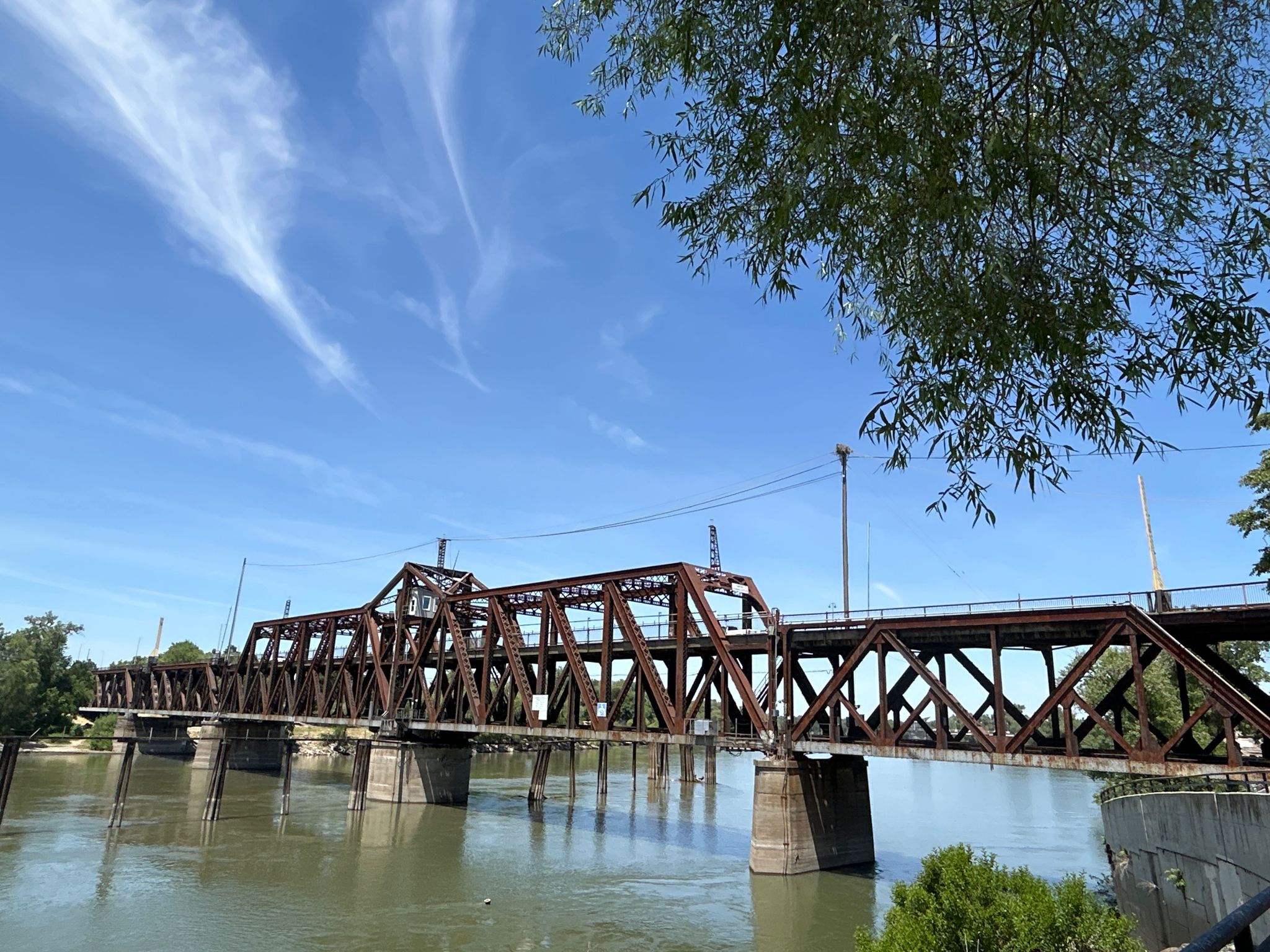A rusty metal truss bridge spans a river with trees and wispy clouds. - Sacramento River Bike Trail – Tower Bridge loop from Pocket + Blue Dolphin