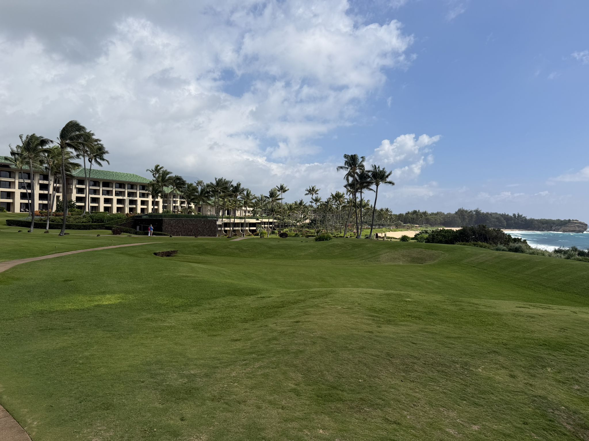 Vast green lawn with buildings, palm trees, and ocean under cloudy sky. - Koloa Heritage Trail