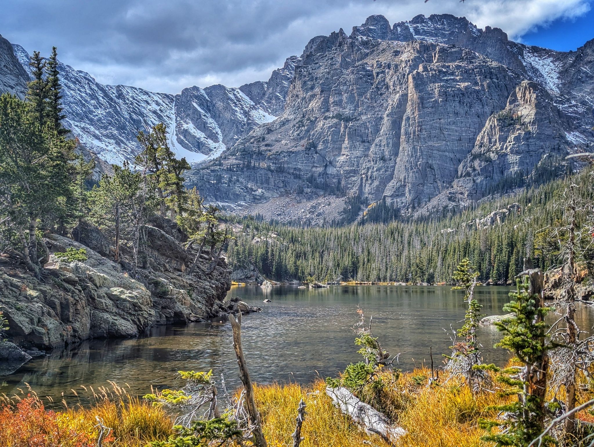 Scenic mountain lake with snow-capped peaks, pine forests, and golden grass under cloudy sky. - Sky Pond Trail