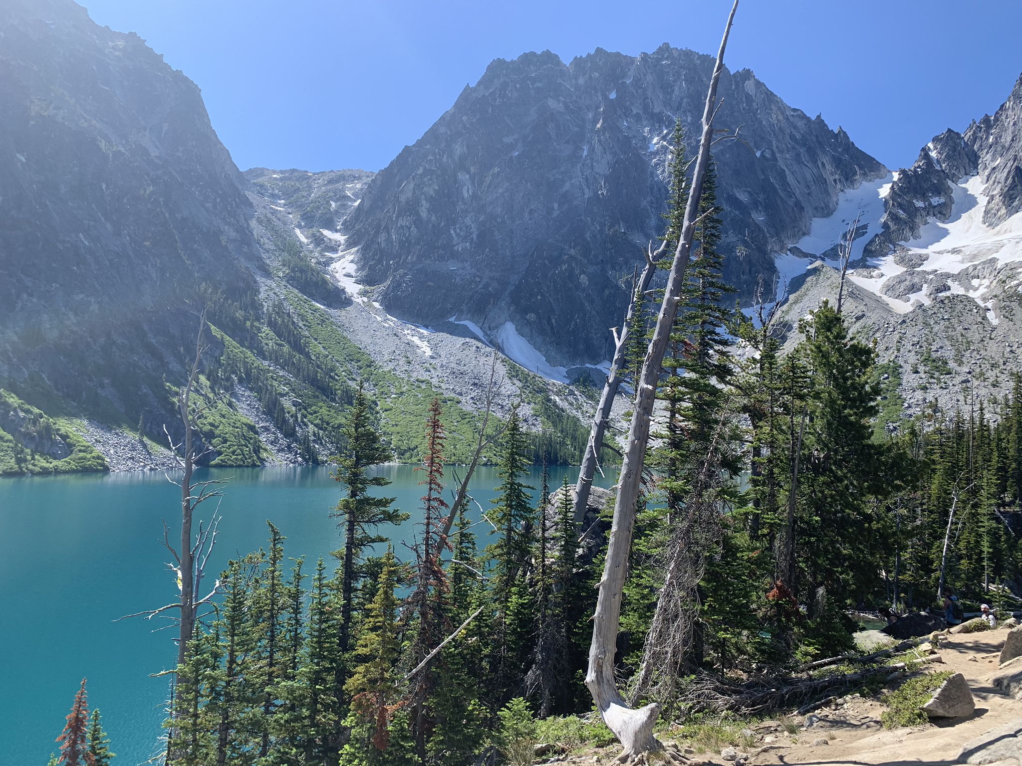Bright turquoise lake with steep mountains, snow patches, and pine trees. - Colchuck Lake Trail