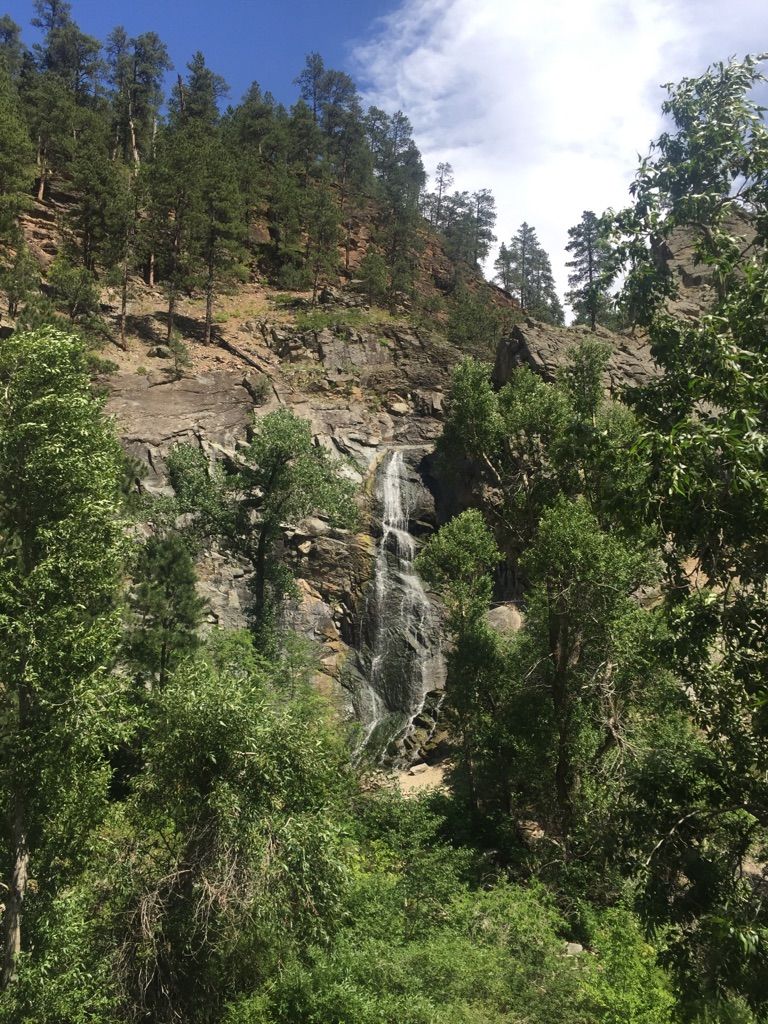 Waterfall flowing down a rocky cliff face, surrounded by green trees. - Spearfish Falls via Spearfish Canyon Scenic Byway