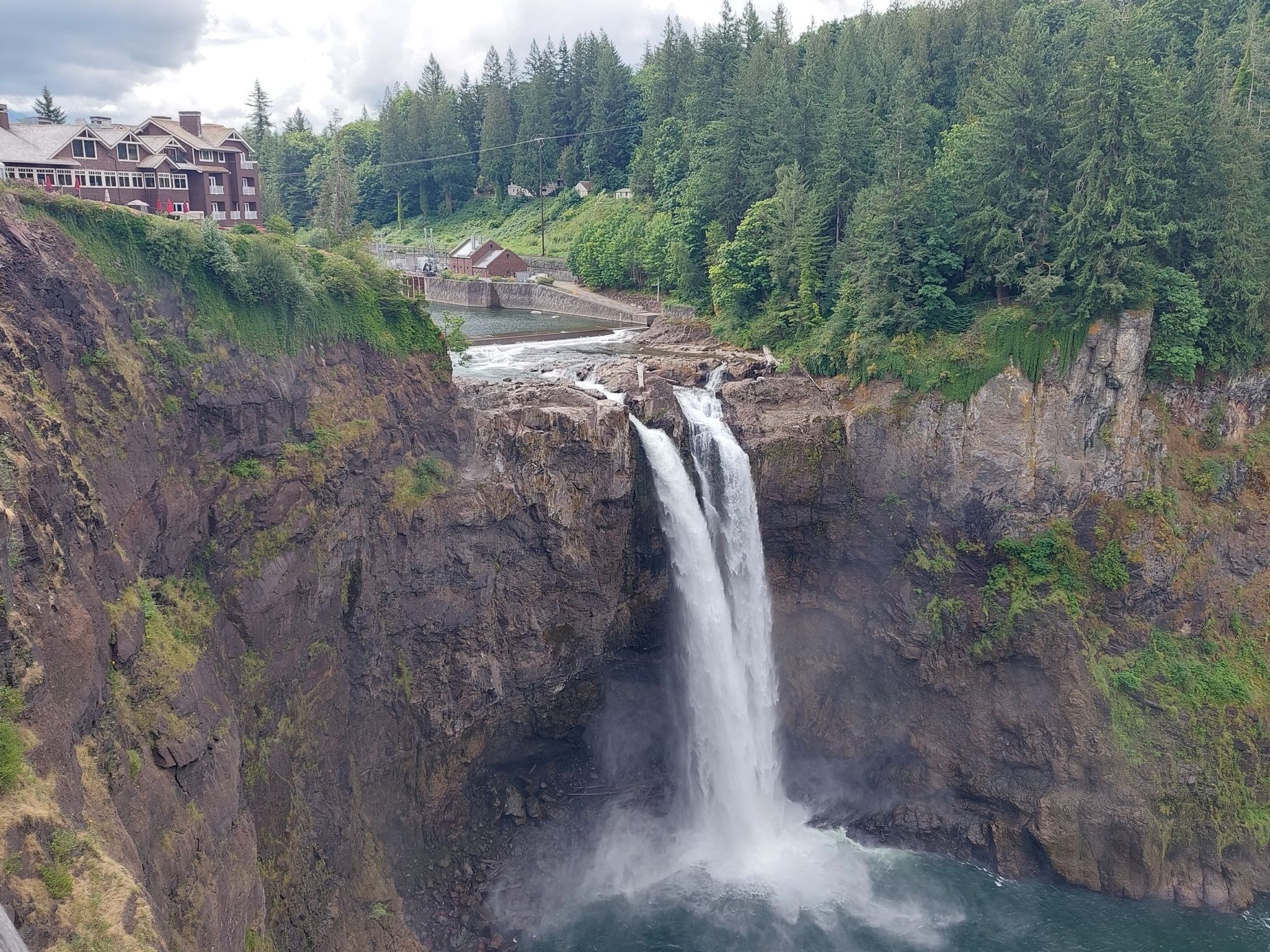 A large waterfall cascades down a cliff, with a building and forest above. - Snoqualmie Falls Trail