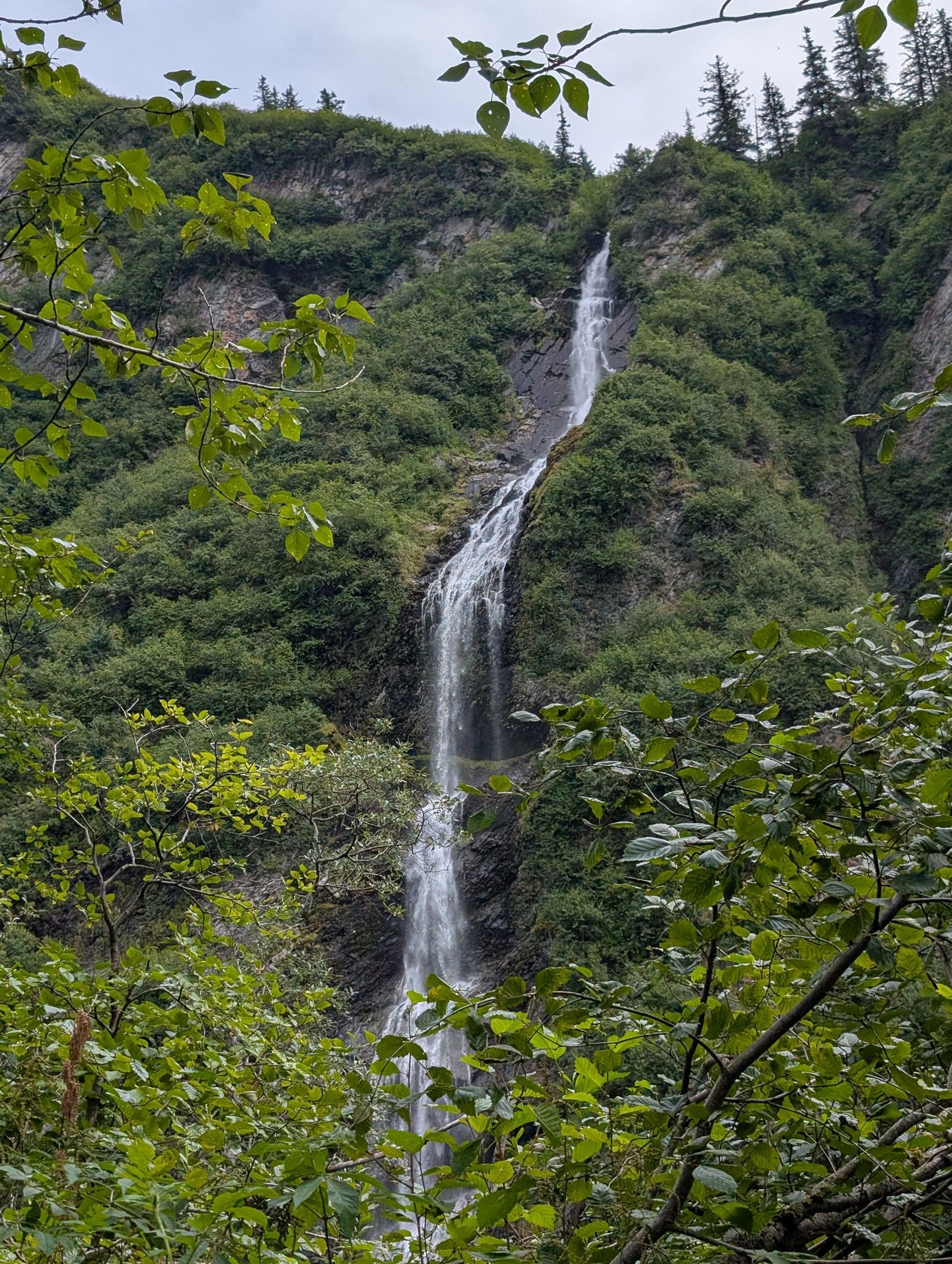 Tall waterfall cascades down a lush green, rocky mountain under an overcast sky. - Goat Trail