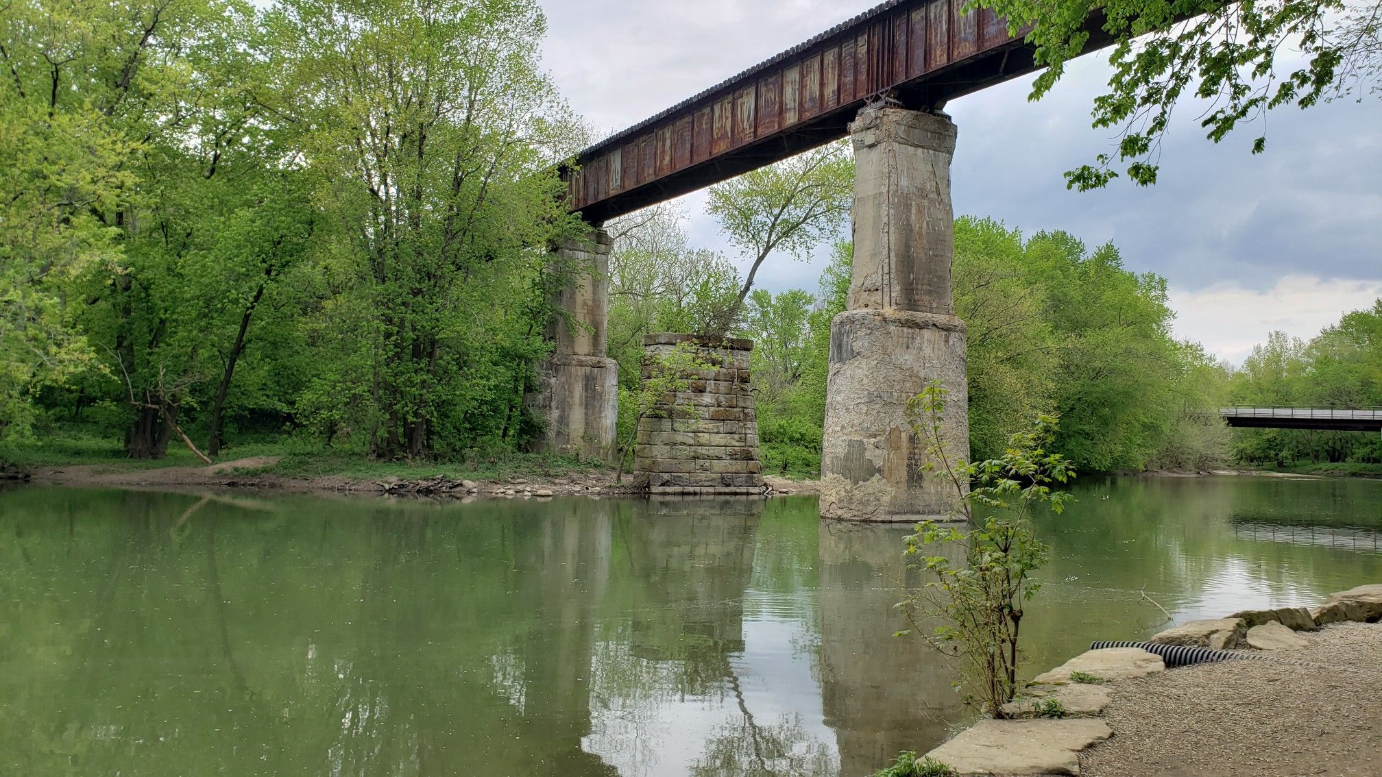 Old railway trestle bridge over a green river surrounded by lush trees. - Ancient Loop
