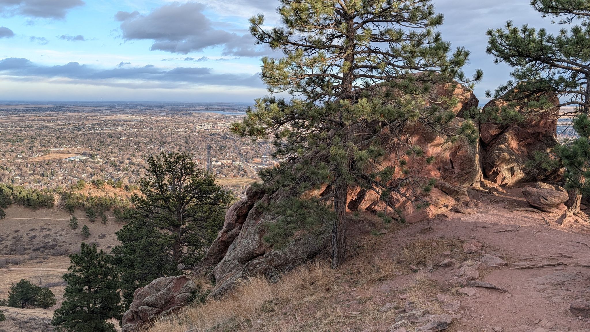 Pine trees and red rocks overlooking a distant town under cloudy sky. - Mount Sanitas Loop