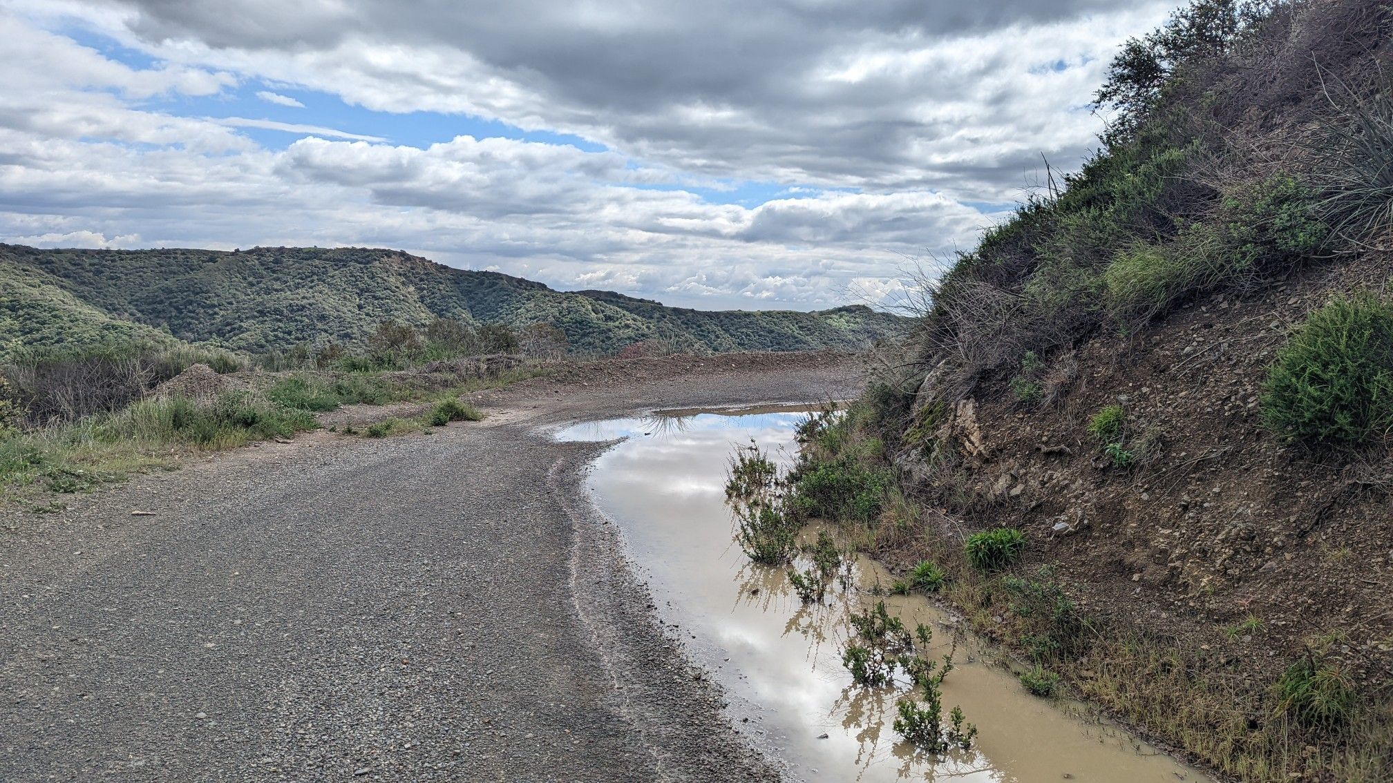 Gravel road next to a muddy stream, green hills under cloudy sky. - San Vicente Mountain Viewpoint – West Mandeville Fire Road loop from Downtown Santa Monica