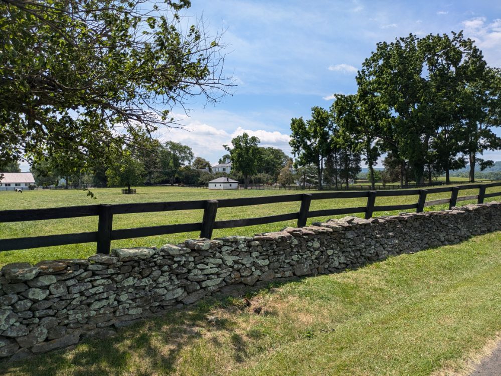 A stone wall and wooden fence border a sunny green field with distant buildings. - Bunker Hill – The Plains loop from Delaplane