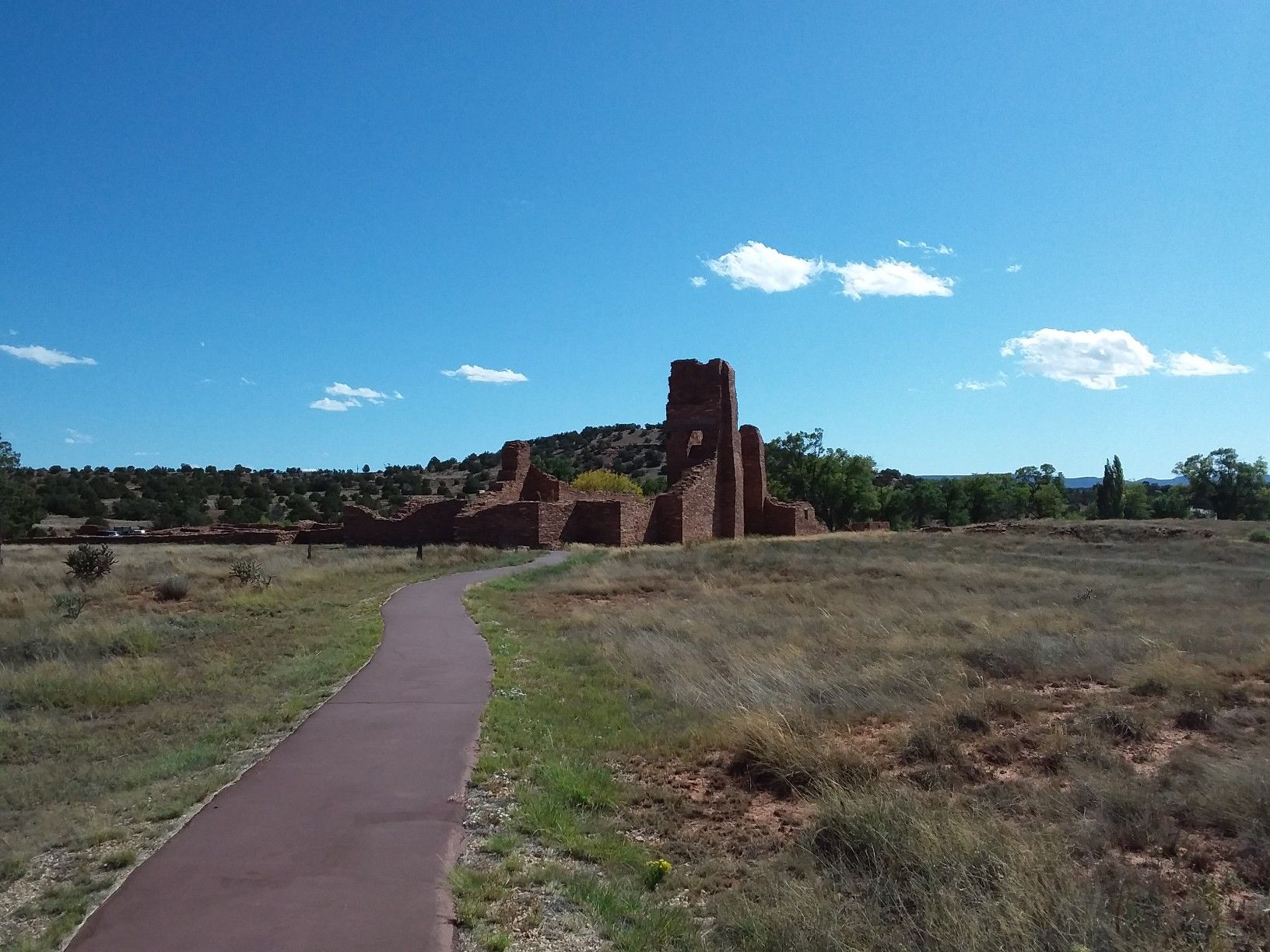 A paved path leads through dry grass towards distant red stone ruins. - Mission Ruins Interpretive Trail
