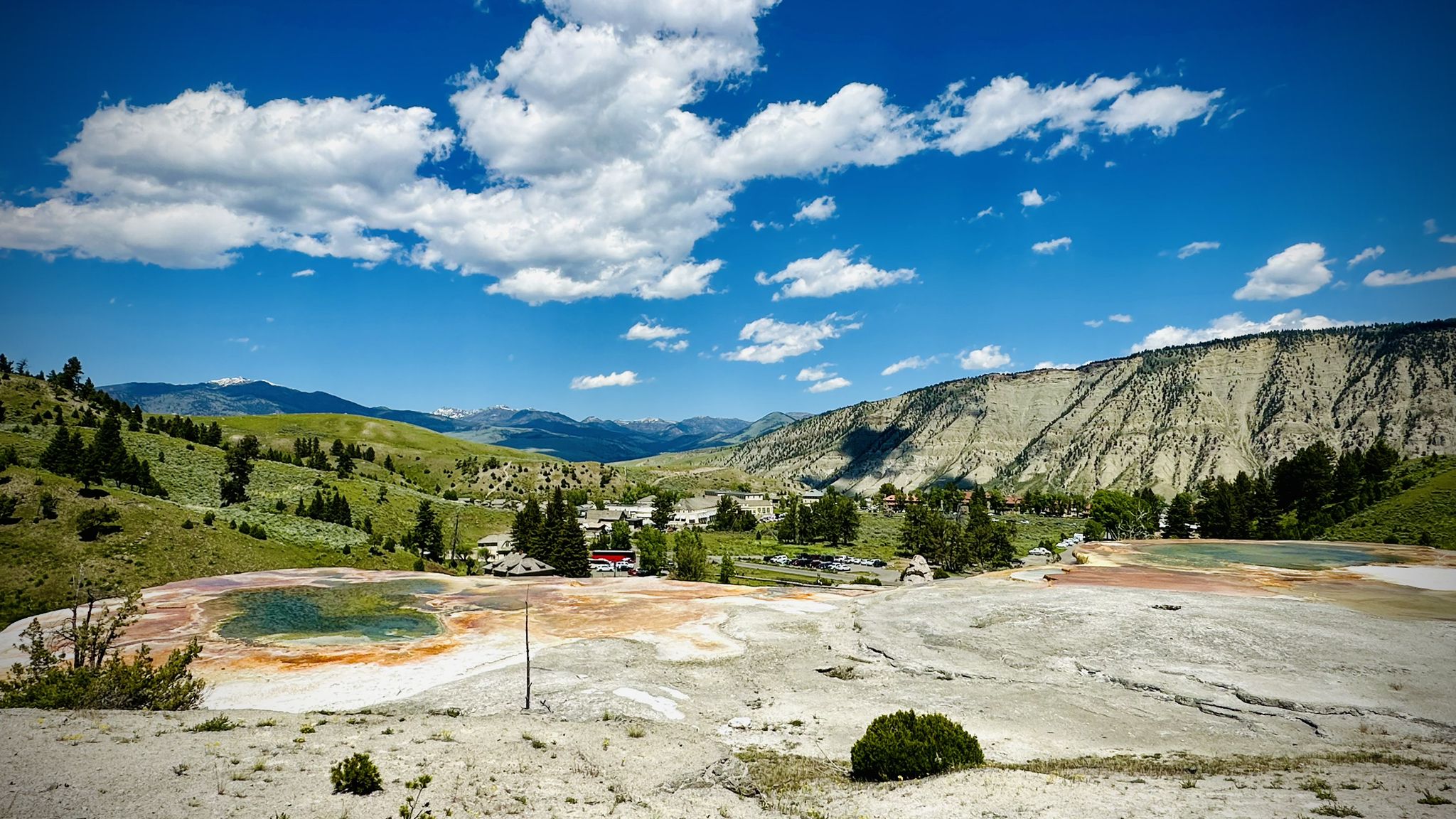 Colorful hot springs and green hills with distant mountains under a bright blue sky. - Palette Springs – Mammoth Hot Springs Terraces loop from Nymph Lake