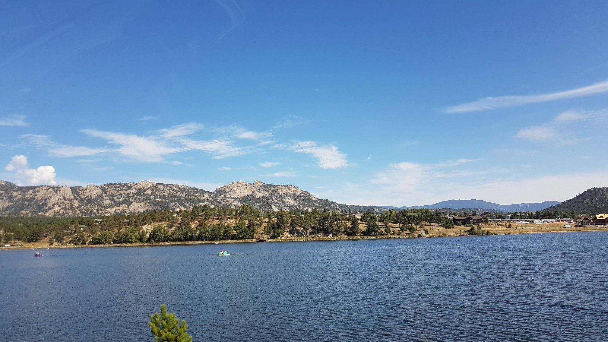 Wide lake view with distant mountains, green trees, and a clear blue sky. - Lake Estes Trail