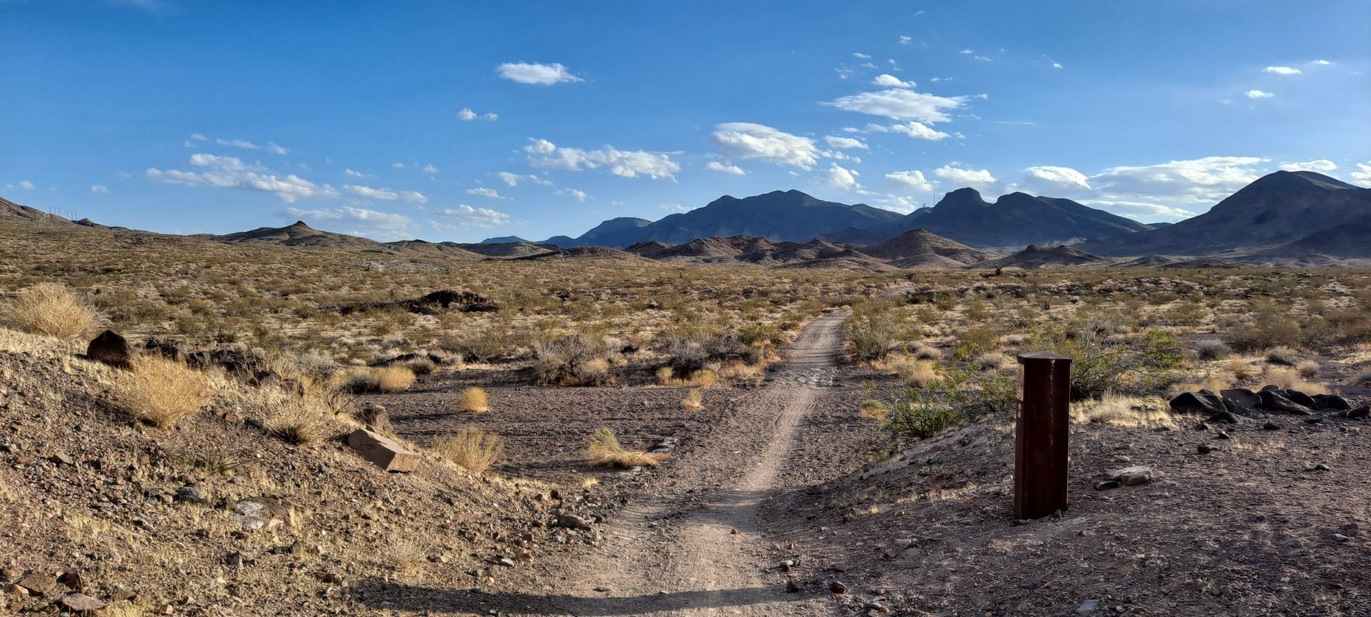 A dirt path winds through a vast, sunny desert landscape with distant mountains. - McCullough and 601 Loop