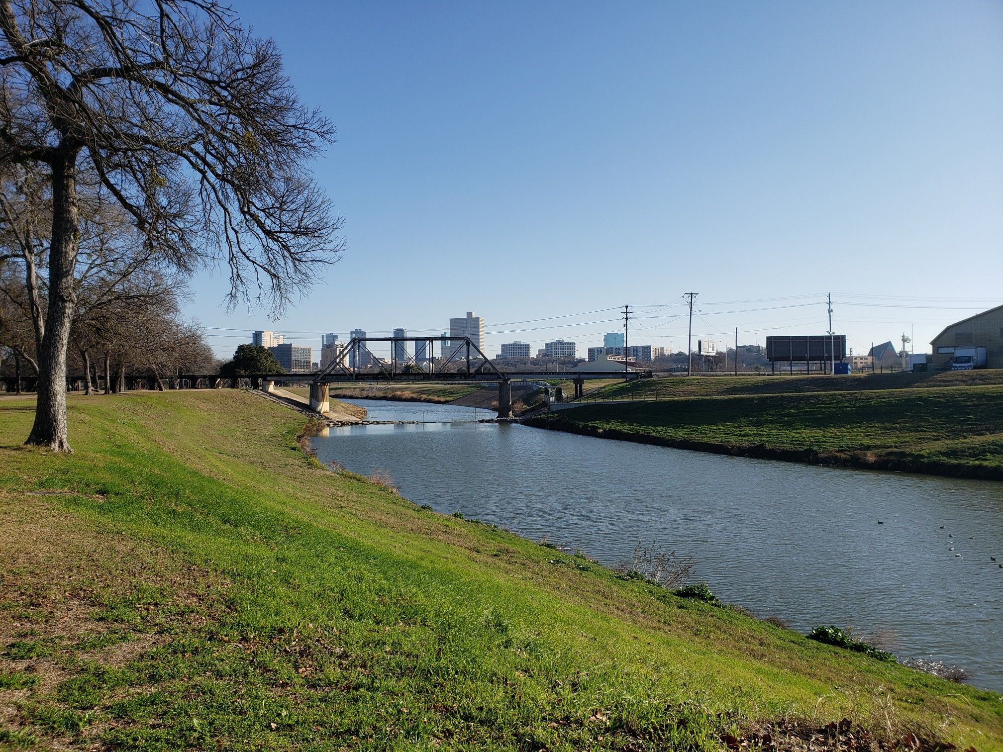 River flowing towards a city skyline with a bridge under a clear sky. - Trinity Trails: Gateway Park Loop