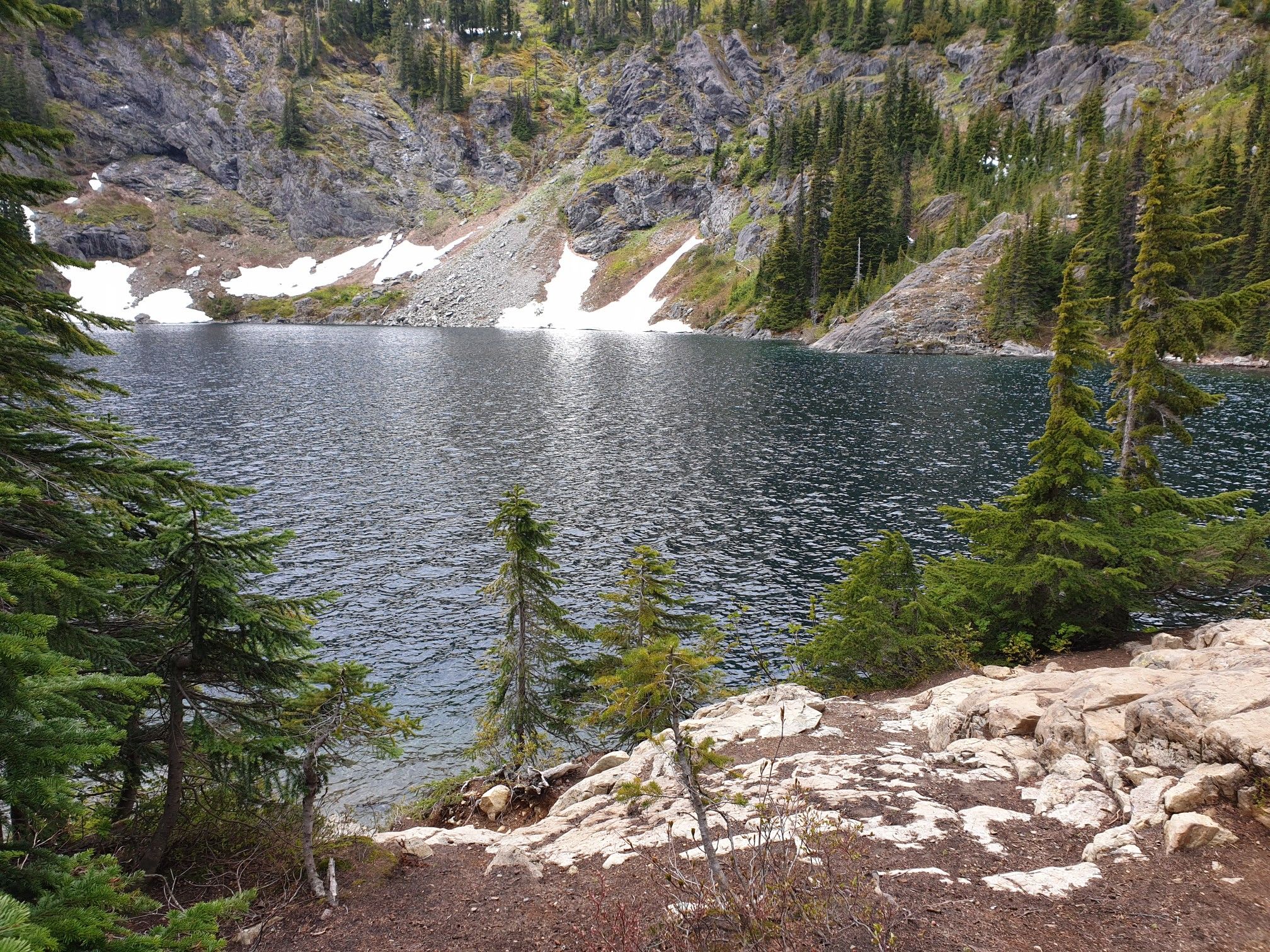 Mountain lake with snow patches, surrounded by pine trees and rocky slopes. - Rachel Lake Trail