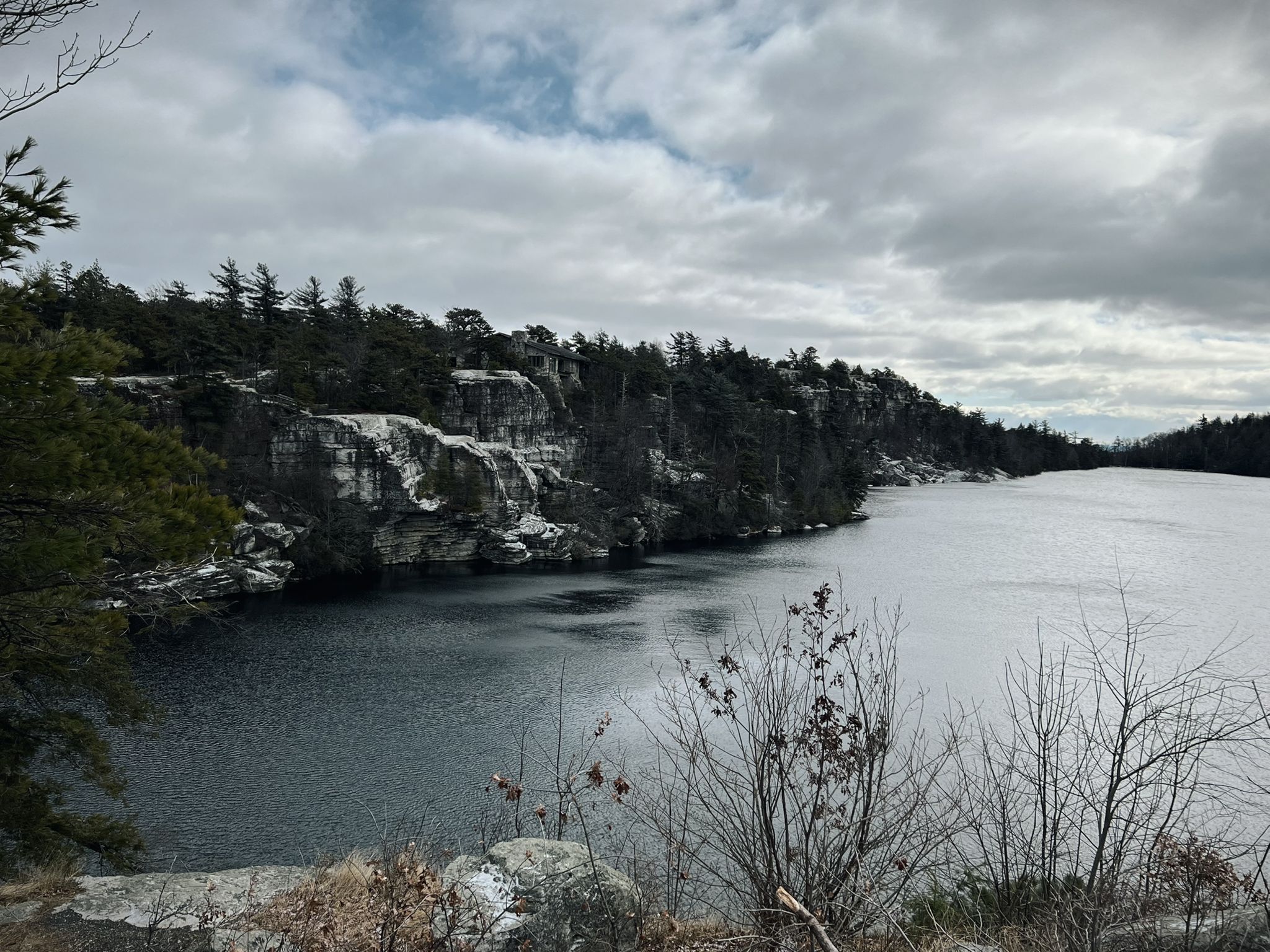 Lake with snow-dusted cliffs and evergreen trees under a cloudy sky. - Lake Minnewaska Carriage Road Loop