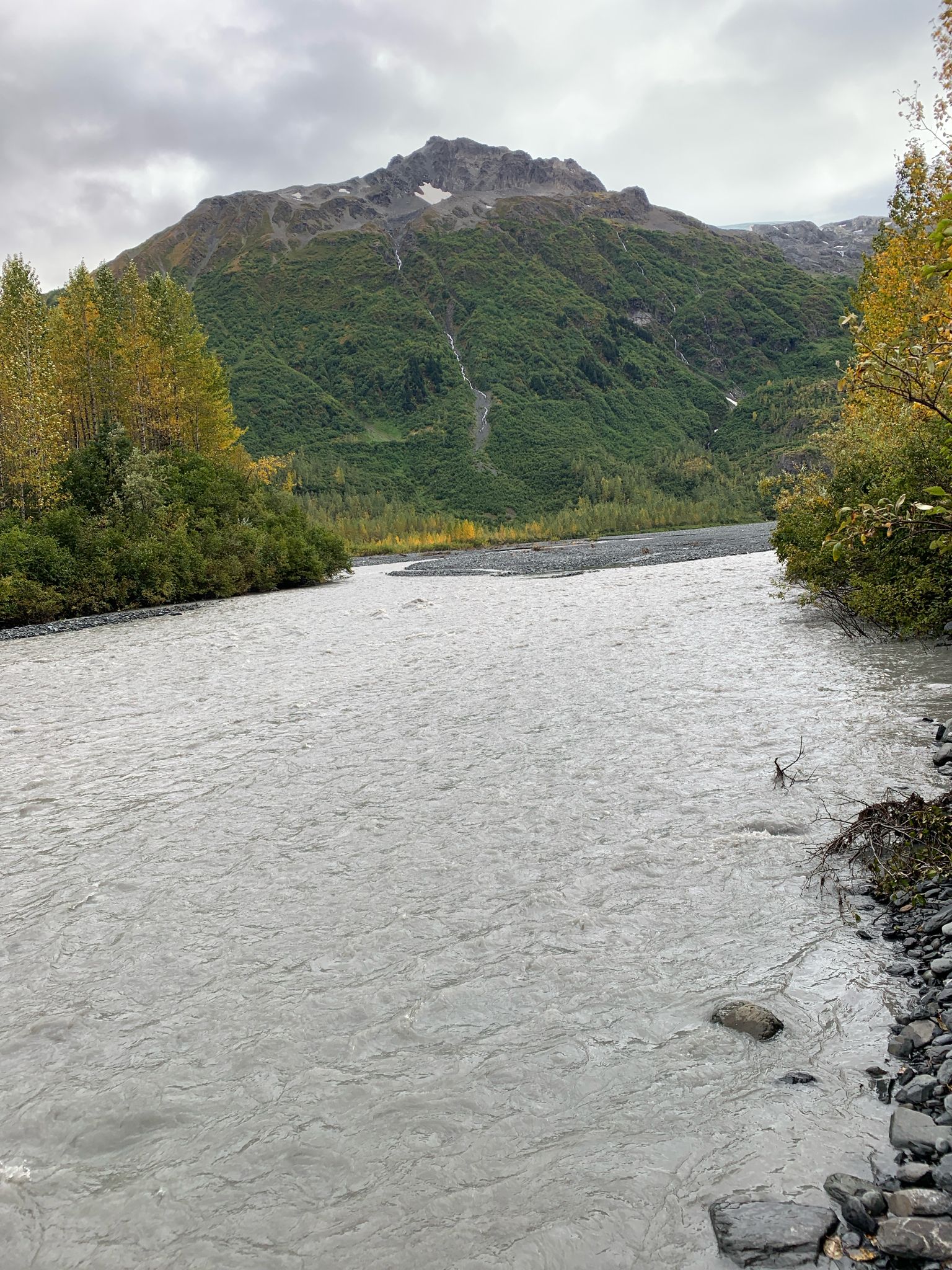 A wide river flows past a green mountain under a cloudy sky. - Glacier View Loop Trail