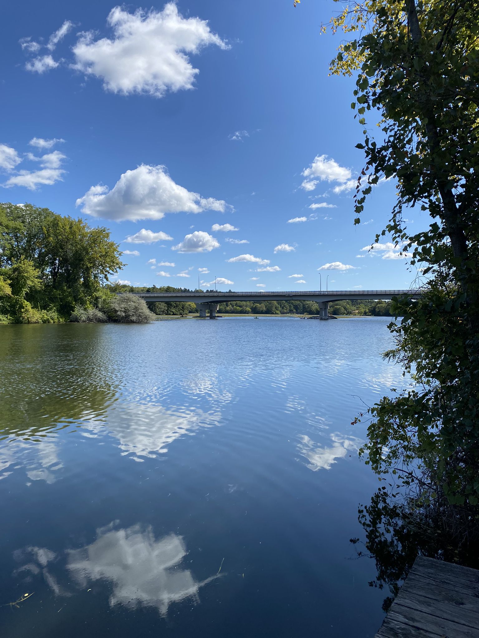 A wide river reflects a blue sky with white clouds, a bridge in the distance. - Parker Mill County Park – Argo Dam Railway Bridge loop from Arbor Hills Nature Area