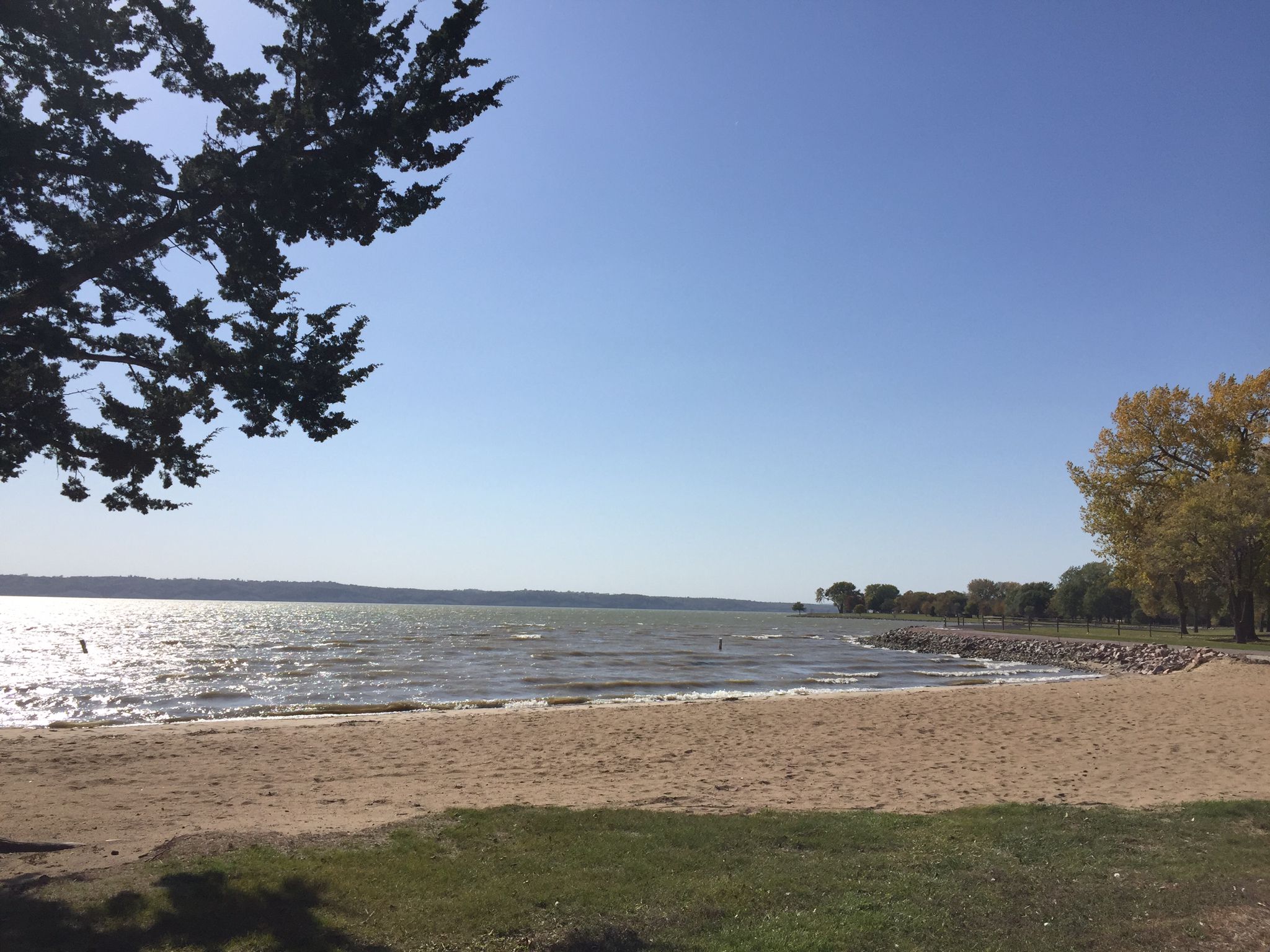 Sunny beach with calm lake water and trees under a clear blue sky. - Lewis and Clark Lake Trail