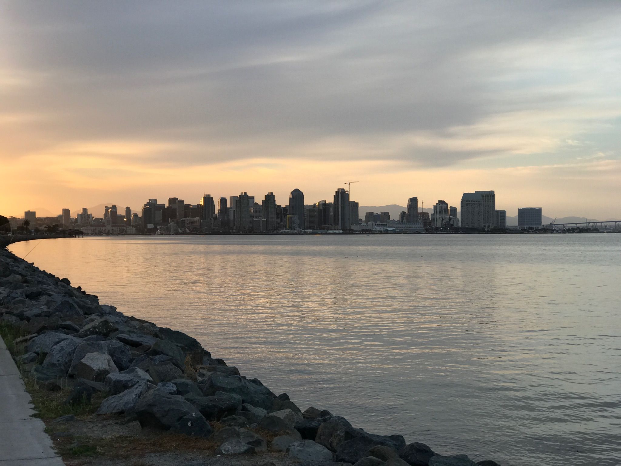 Distant city skyline across calm water with a rocky foreground under an overcast sky. - Spanish Landing Monument – Spanish Landing Park loop from Point Loma