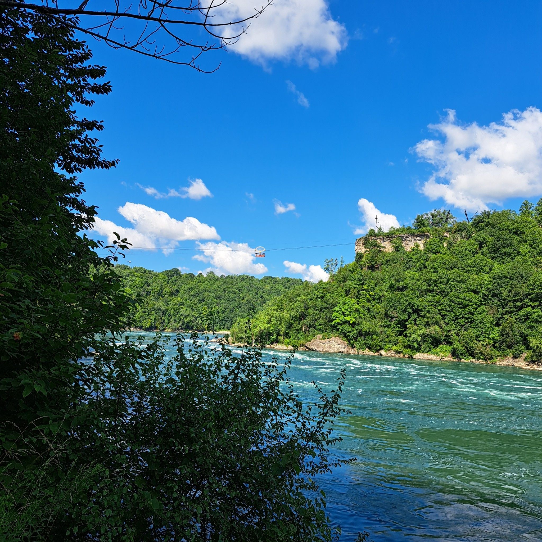 Wide river flowing between lush green hills under a blue sky with clouds. - Niagara Gorge Rim and Whirlpool Rapids Trail