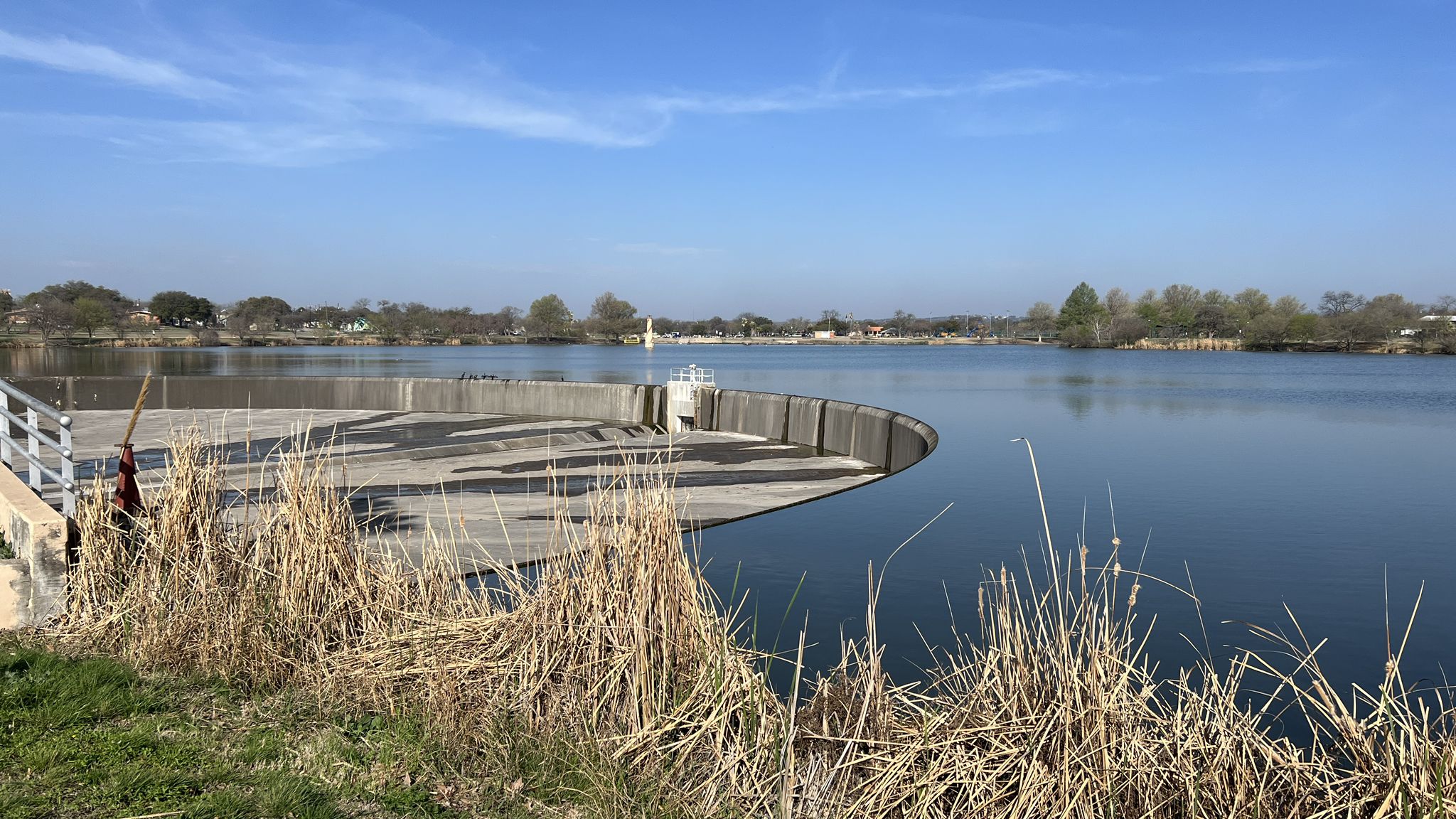 Bright daylight over a calm lake with a curved concrete dam and dry foreground grass. - Woodlawn Lake Loop