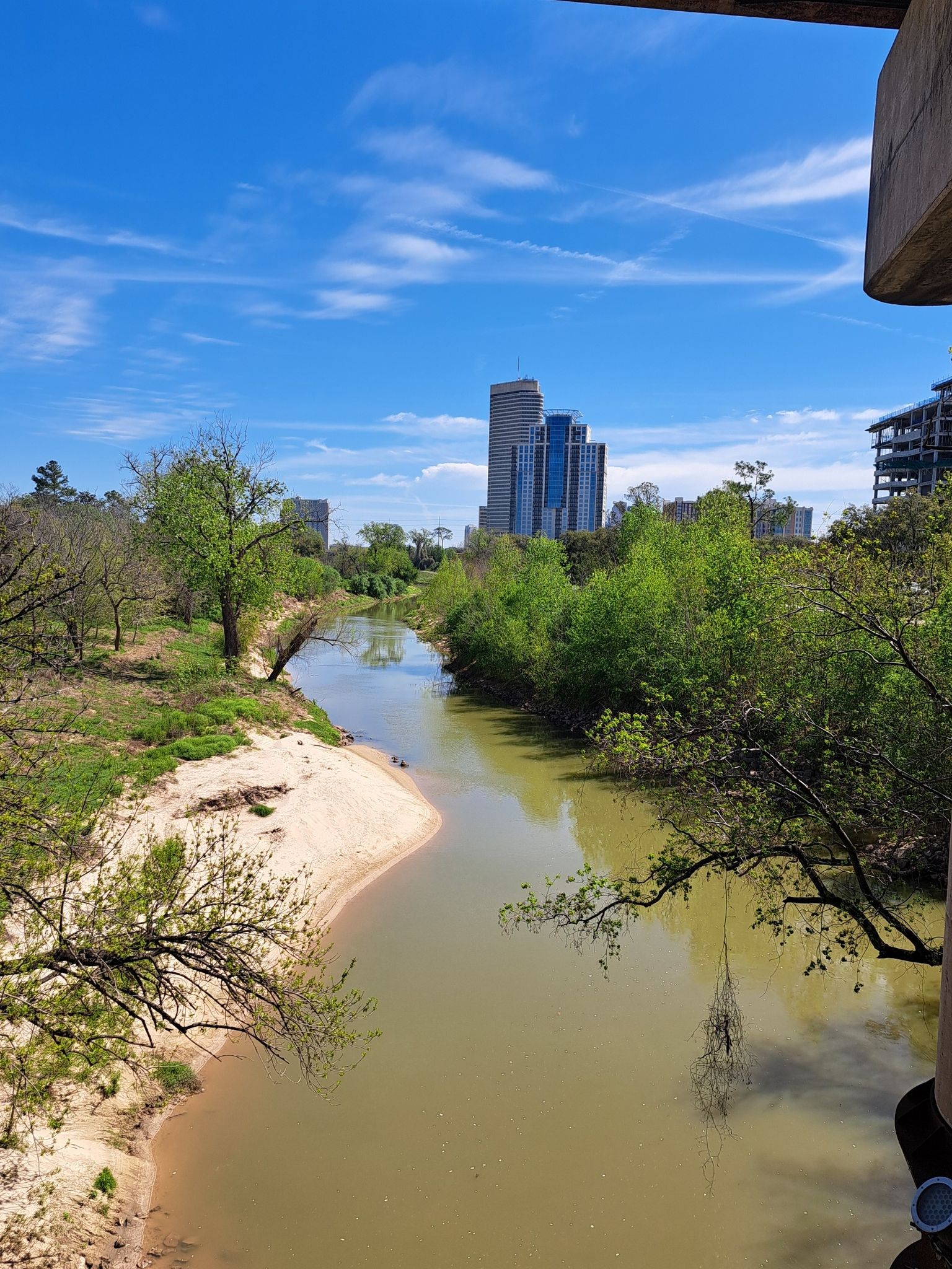 Winding river with a sandy bank, green trees, and distant city buildings. - Buffalo Bayou Park loop from Theater District (eastbound)