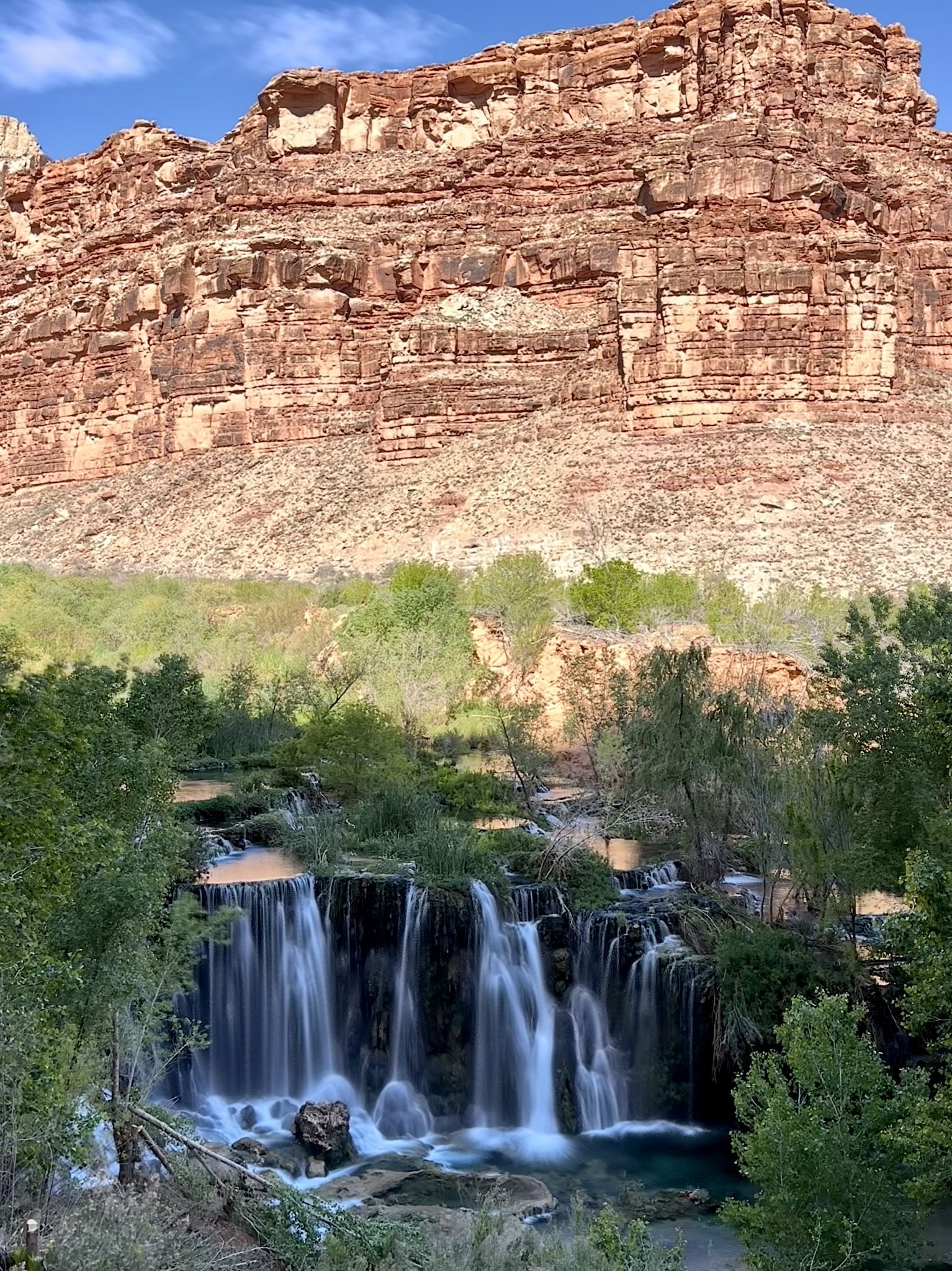 Multi-tiered waterfalls flow below massive red rock cliffs under a bright sky. - Supai Village loop from Supai