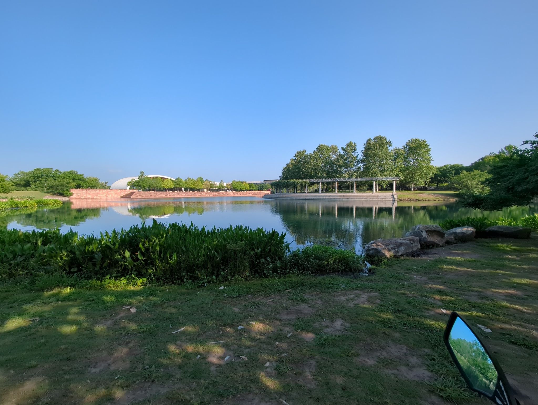 A calm pond reflects a distant circular structure under a clear blue sky. - Mueller Lake Park Trail