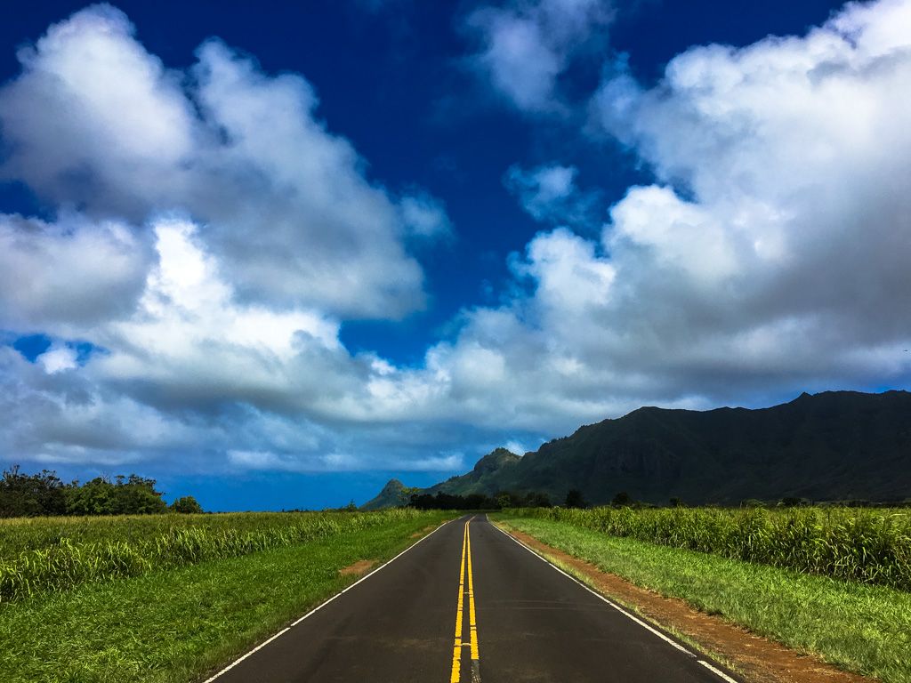A paved road leads through green fields towards mountains under a dramatic sky. - The Tree Tunnel – Kauai Plantation Railway loop from Kauaʻi Plantation Railway