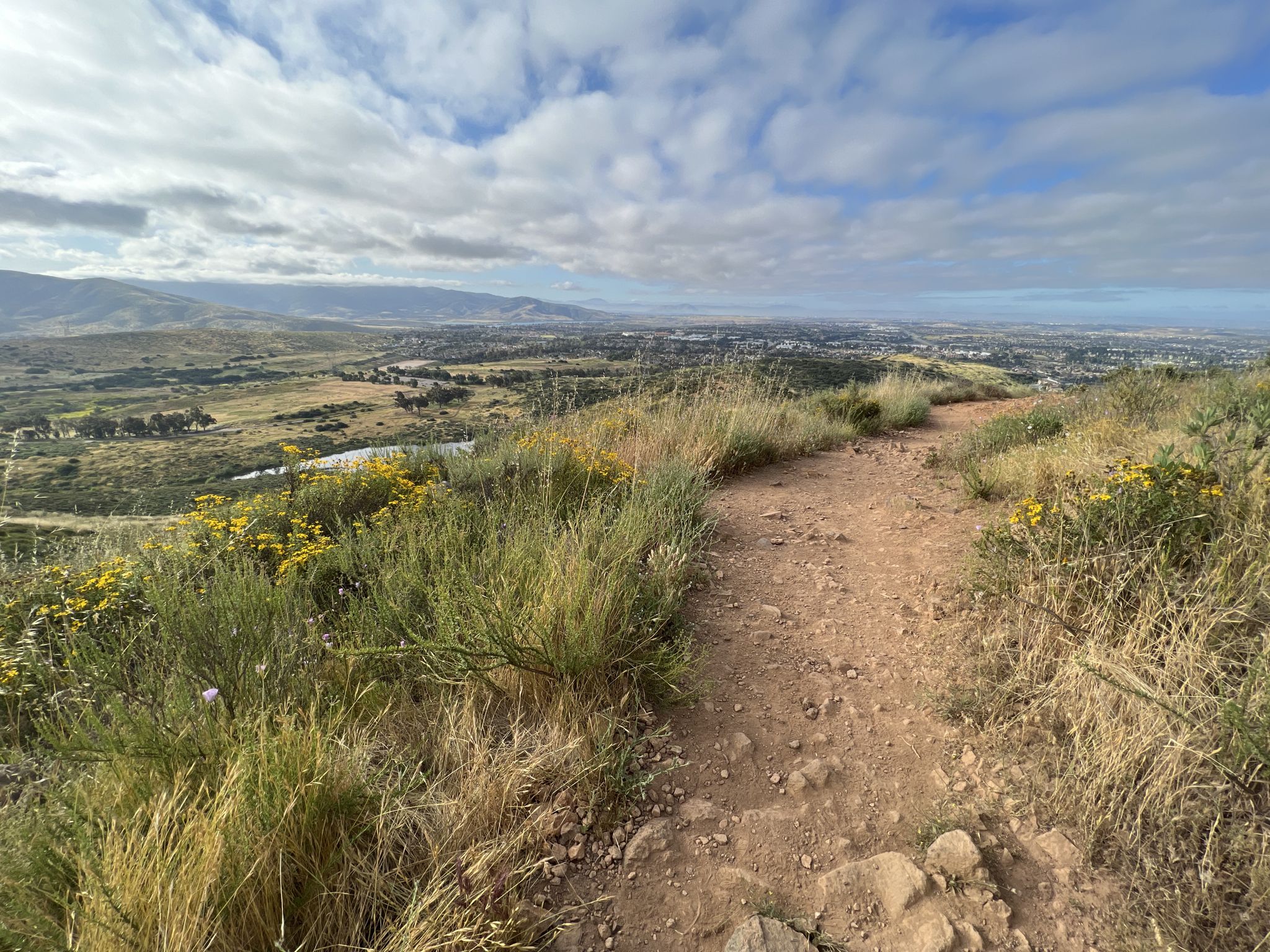 Dirt trail on a grassy hill overlooking a city and lake under a cloudy sky. - Mother Miguel Mountain via Rock House Trail