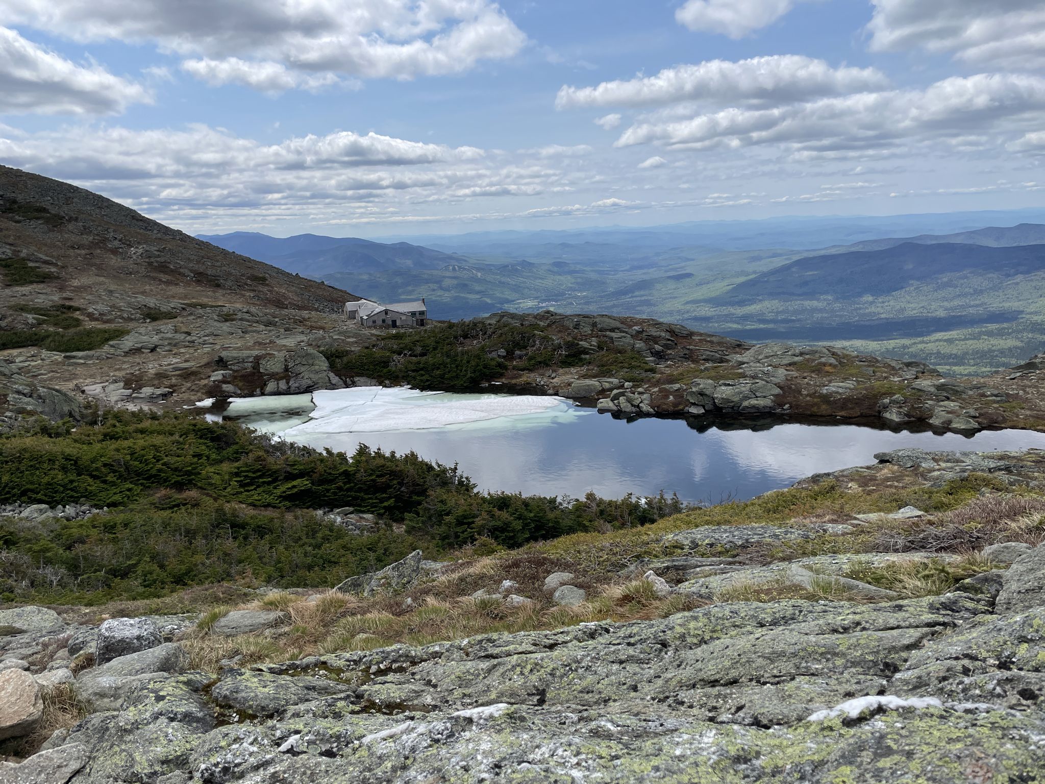 Partially frozen mountain lake with a distant building under a cloudy sky. - Presidential Traverse trail run
