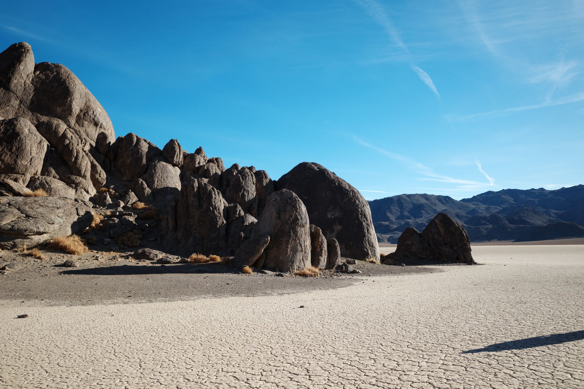 Large dark boulders on a vast cracked desert floor under a clear blue sky. - Racetrack Valley Road