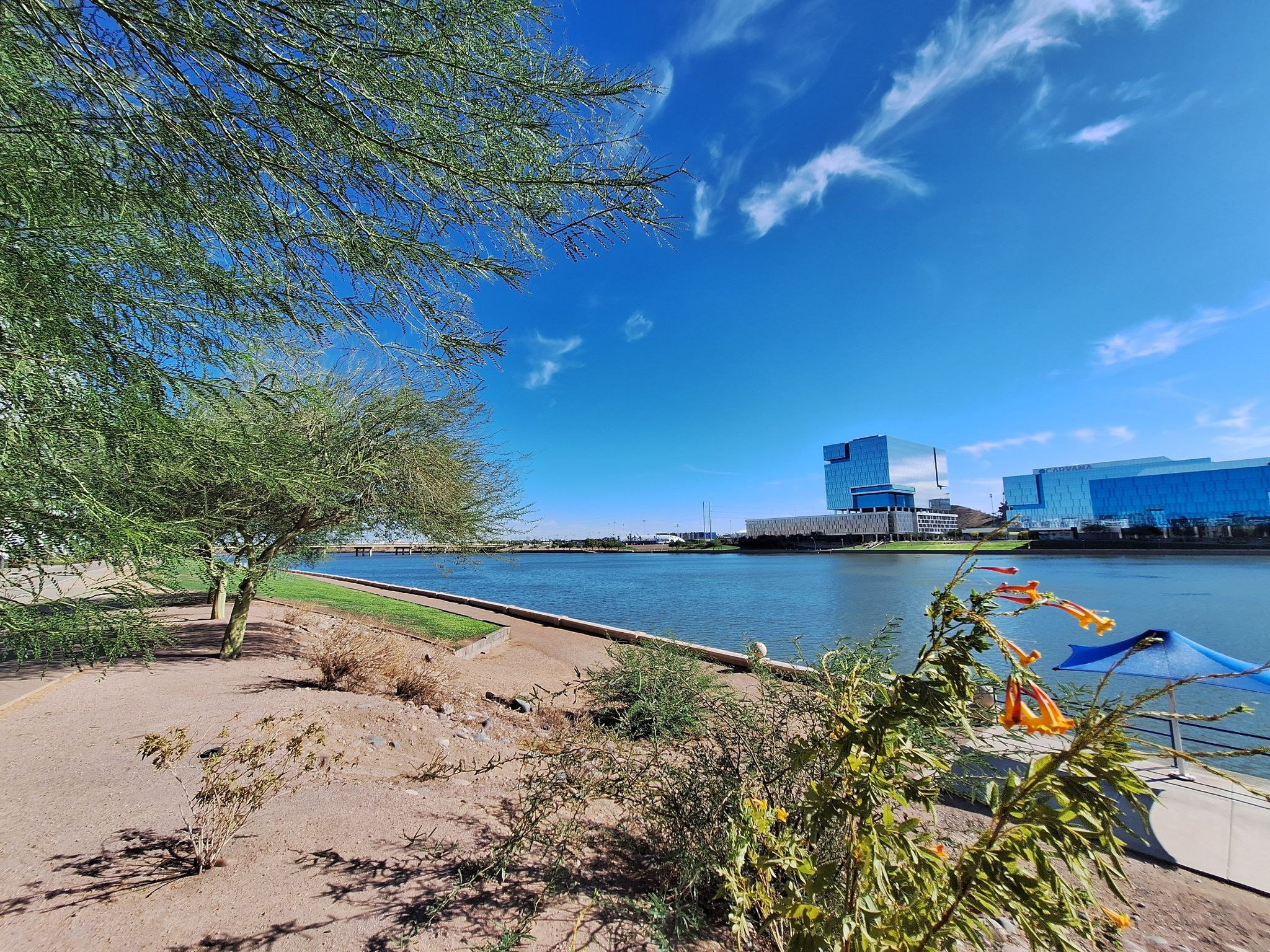 A wide river with sandy banks, trees, and modern buildings under blue sky. - Scottsdale Shea Medical Center & Tempe Town Lake loop