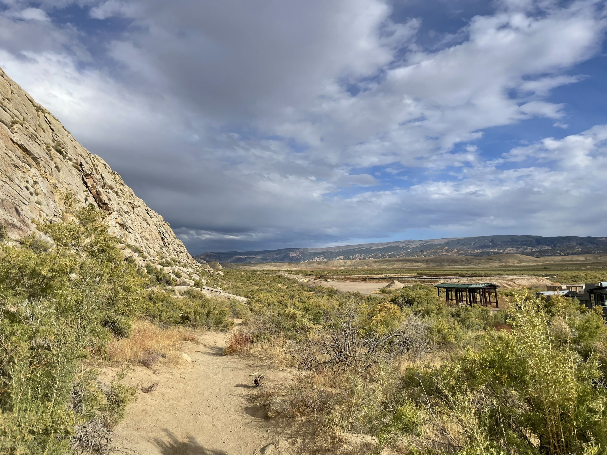 Dirt path leads past a large rock formation under a cloudy sky. - Fossil Discovery Trail – Dinosaur National Monument
