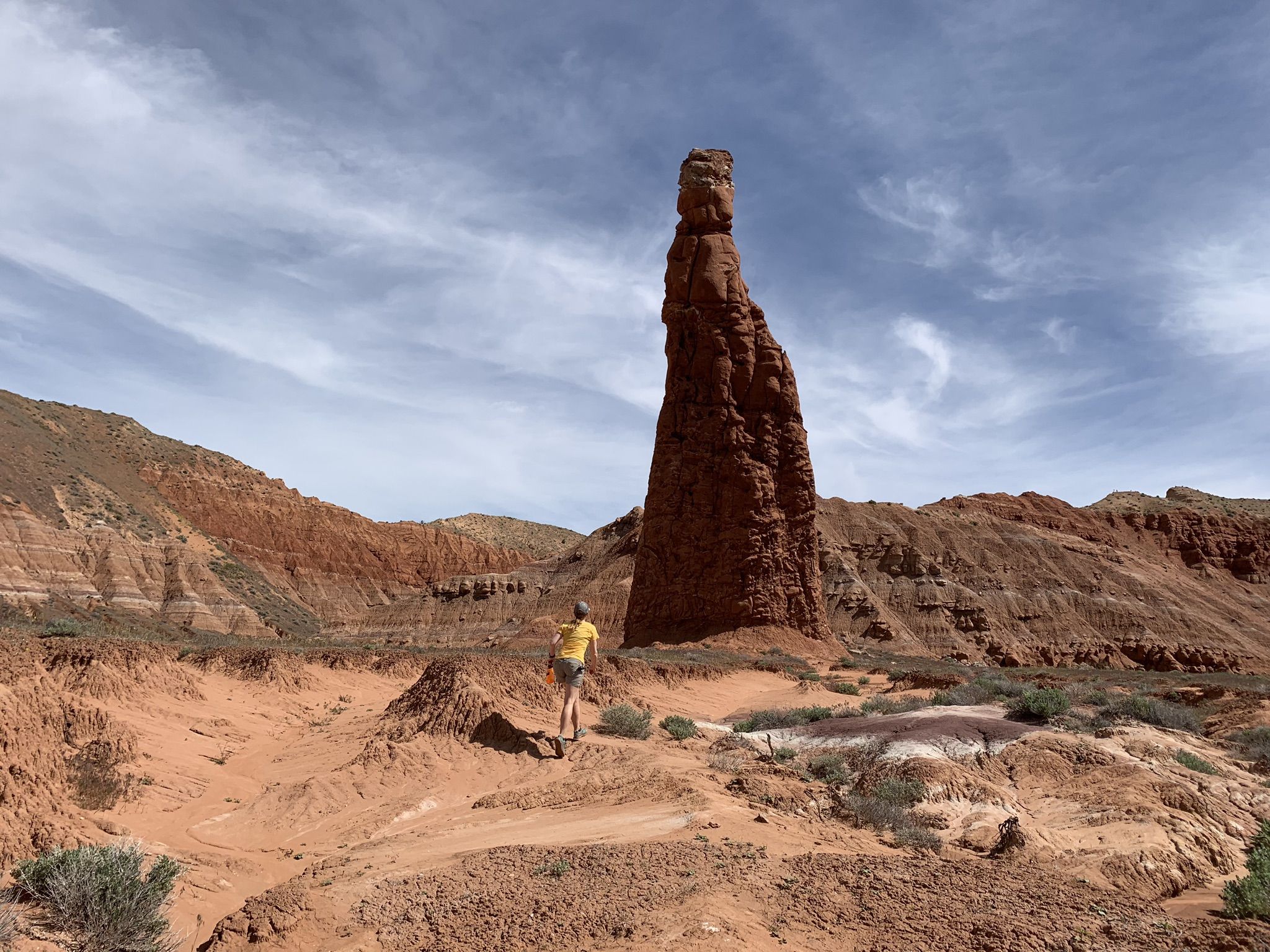 A person stands near a tall, slender red rock formation in a desert landscape. - Trail To Aetschi Baetschi Tower