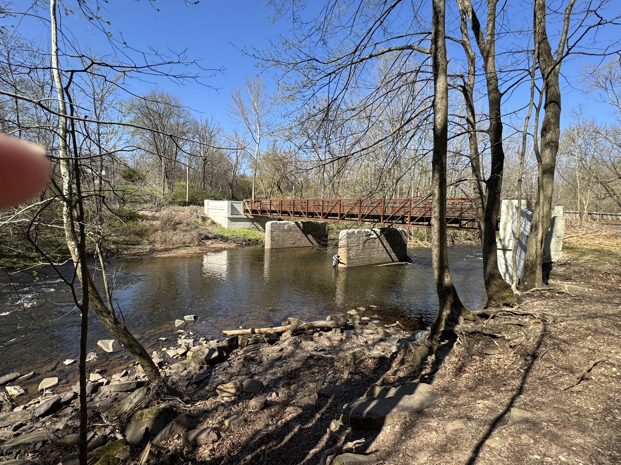 A river flows under a brown bridge, surrounded by bare trees in daylight. - Fern Avenue Trail