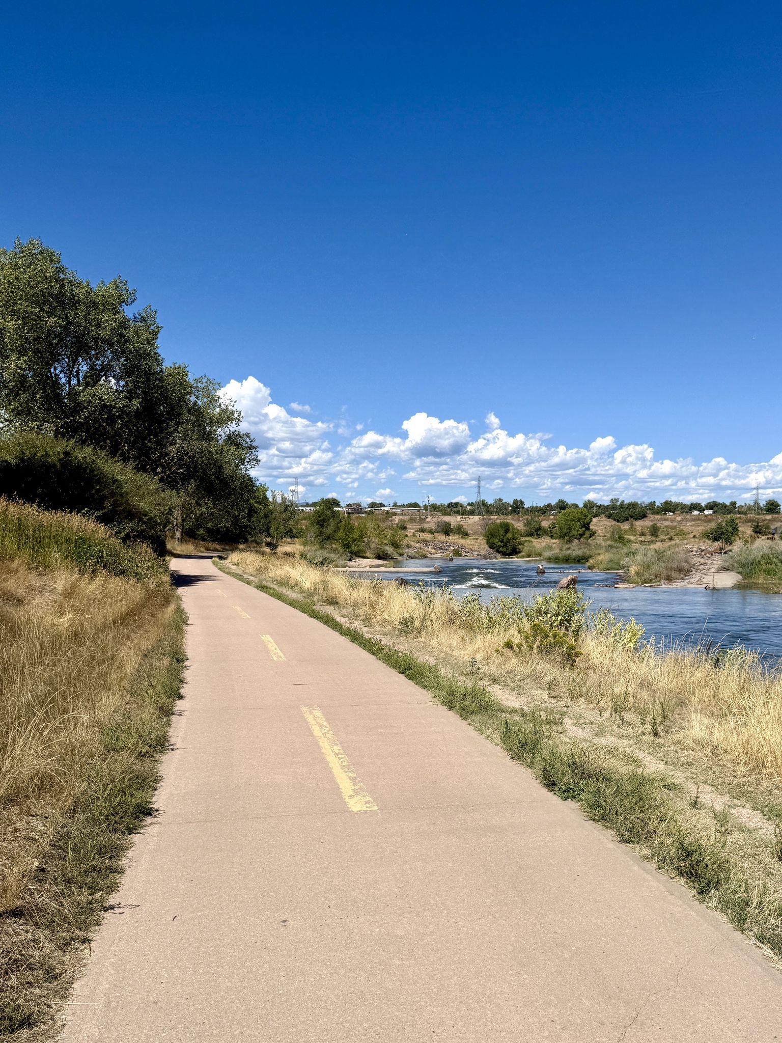 A paved path alongside a river with rapids under a bright blue sky. - Centennial Pond Sunset View loop from Sheridan