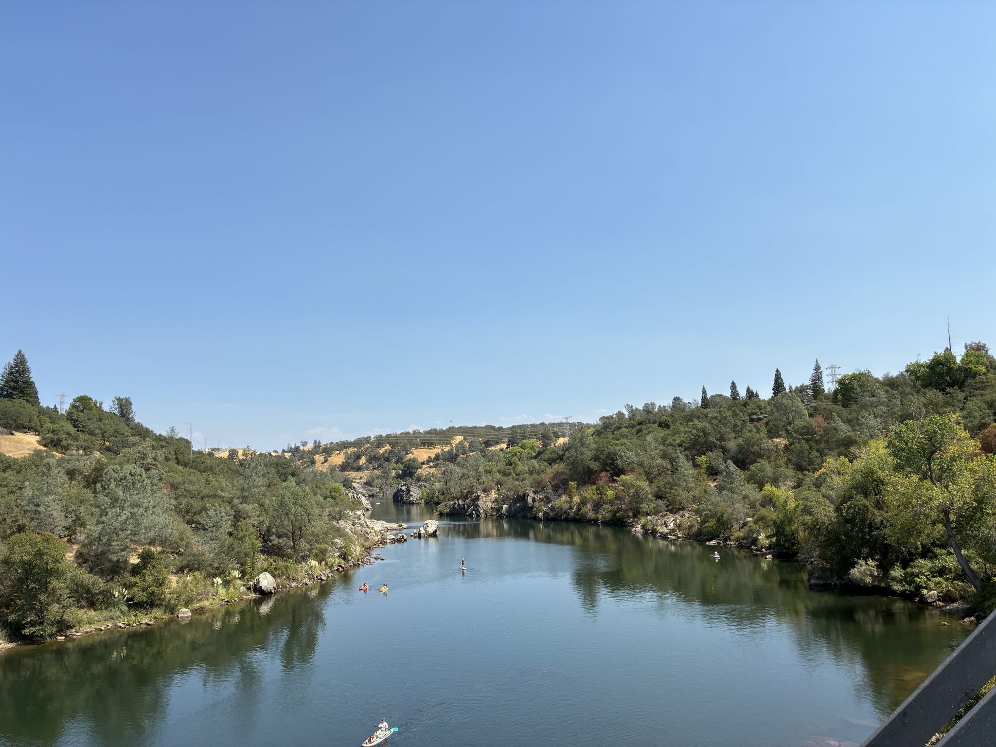 Wide river with kayakers, flowing between wooded hills under a bright blue sky. - American River Bike Trail