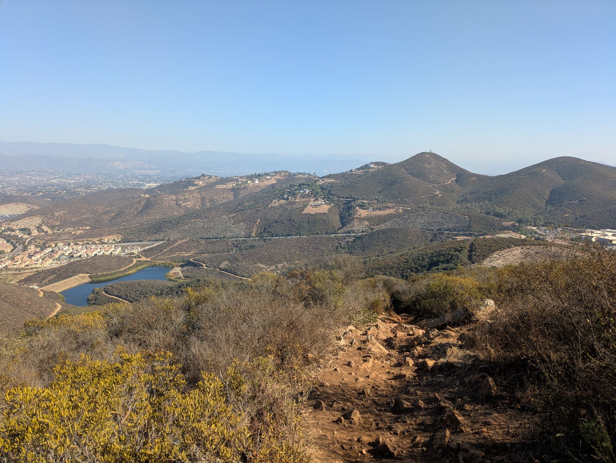 A dirt trail overlooks hills, a small lake, and distant ocean on a clear day. - Double Peak Summit View – Double Peak loop from San Marcos