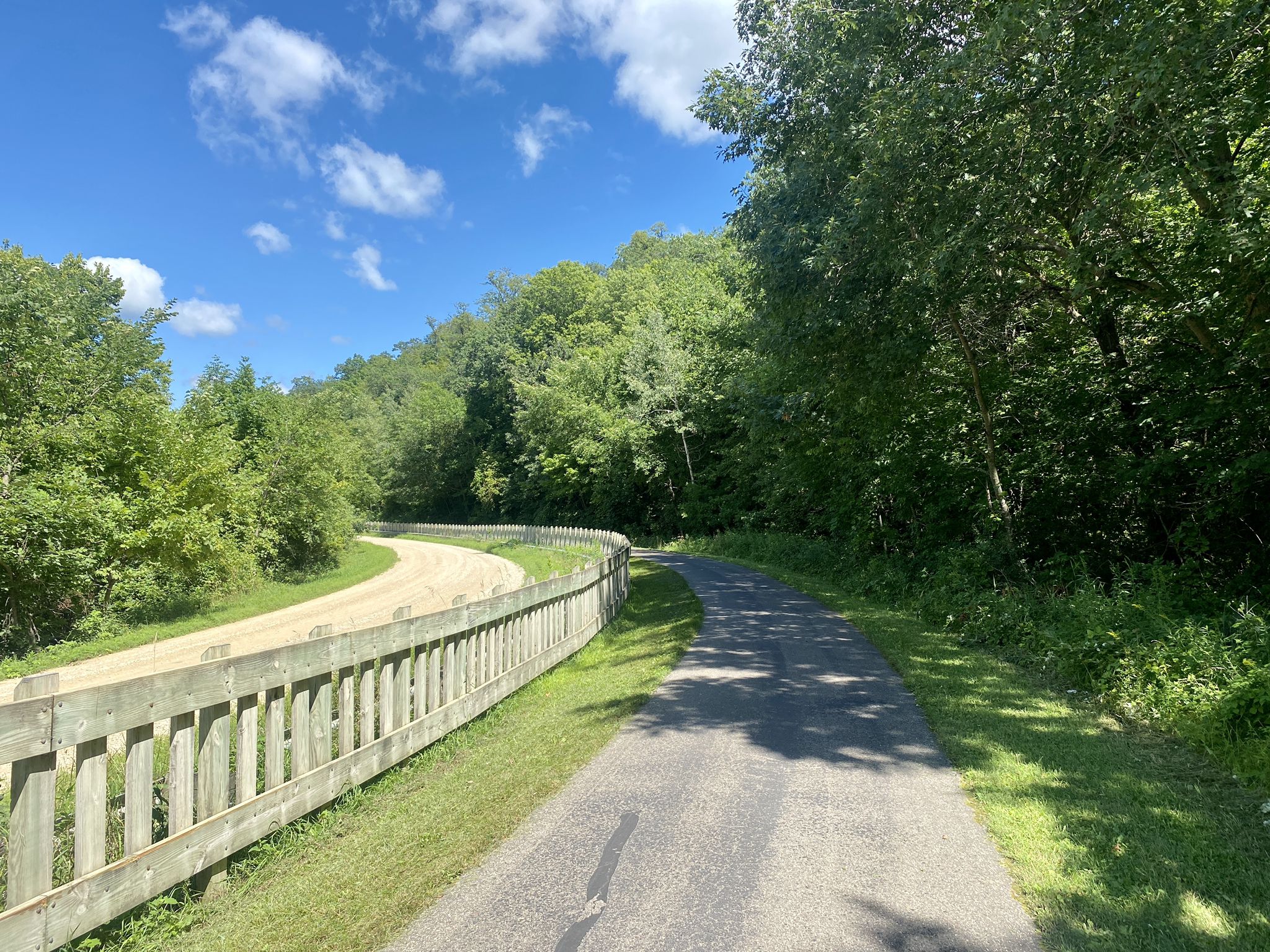 A paved path and dirt path with a fence, surrounded by green trees. - Welch Village – M80 - Train Wreck Site loop from Welch