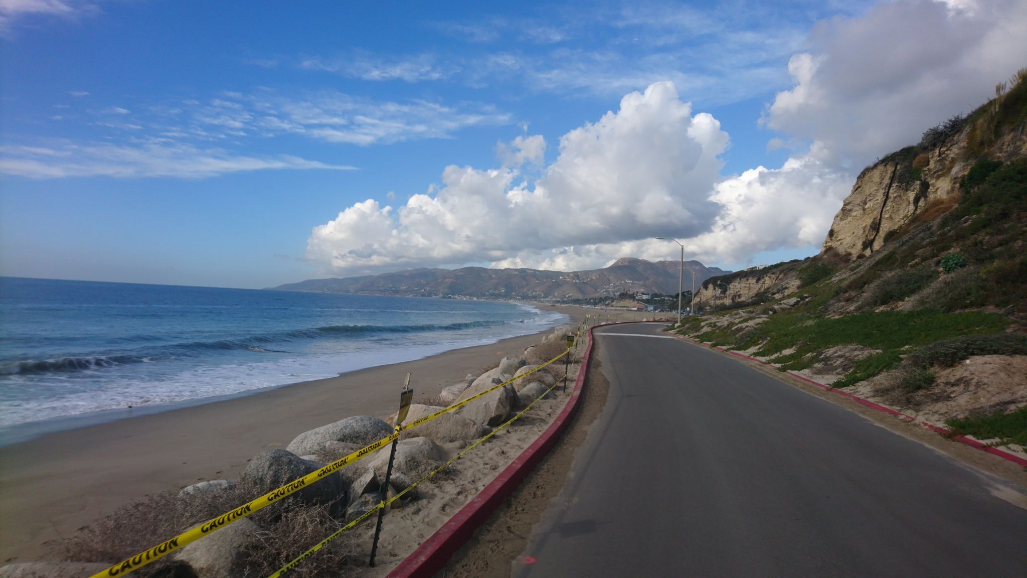 Coastal road beside a beach with waves, cliffs, and mountains under a cloudy sky. - Westward Beach – Point Dume loop from Zuma Beach