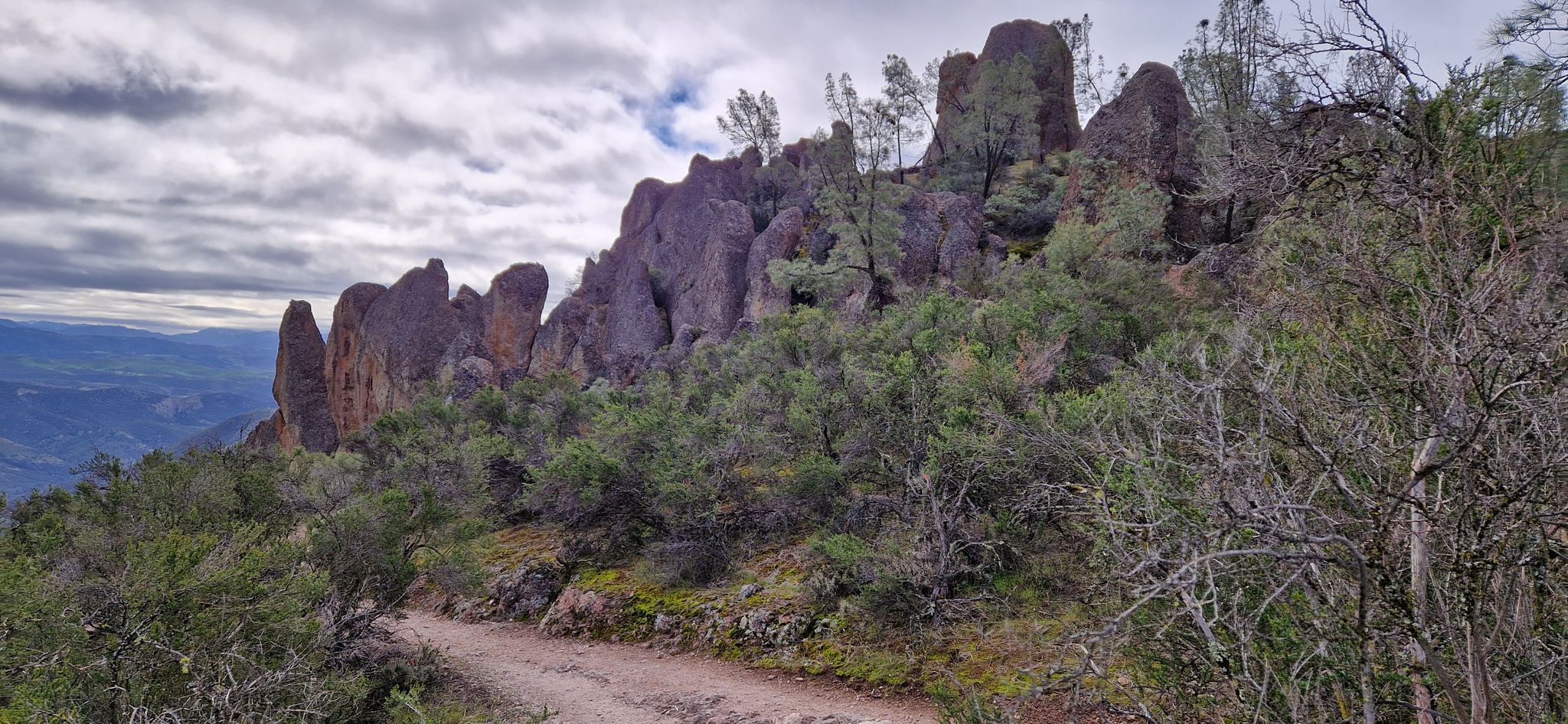 Jagged rock formations rise above a dirt path under an overcast sky. - Bear Gulch Cave – Condor Gulch Trail loop from Jawbone Parking Area