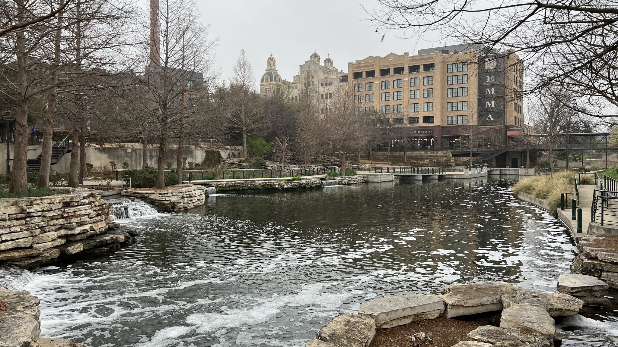 Overcast view of a river with small waterfalls and distant buildings. - River Walk – San Antonio River Walk loop from Trinity University