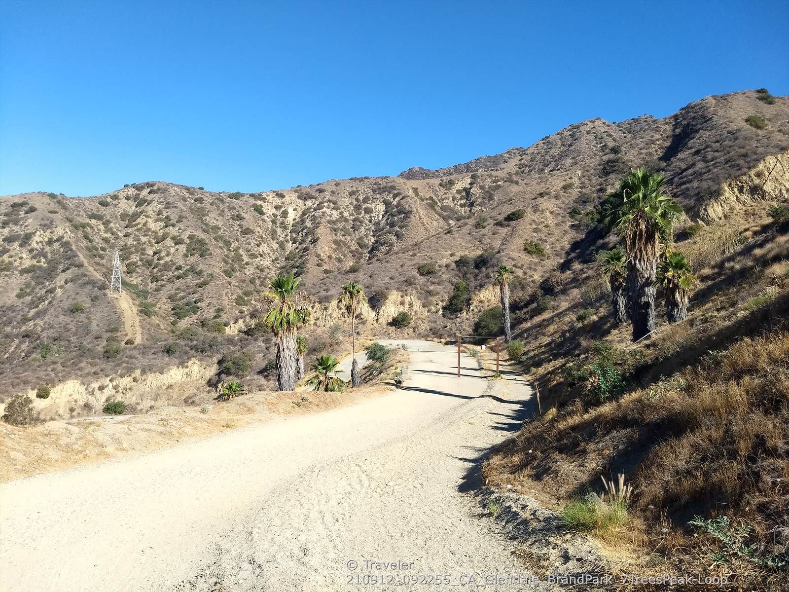 A wide dirt road ascends between dry, scrub-covered hills and palm trees. - Top of Switchbacks — Overlook – Brand Library & Park loop from Herbert Hoover High School