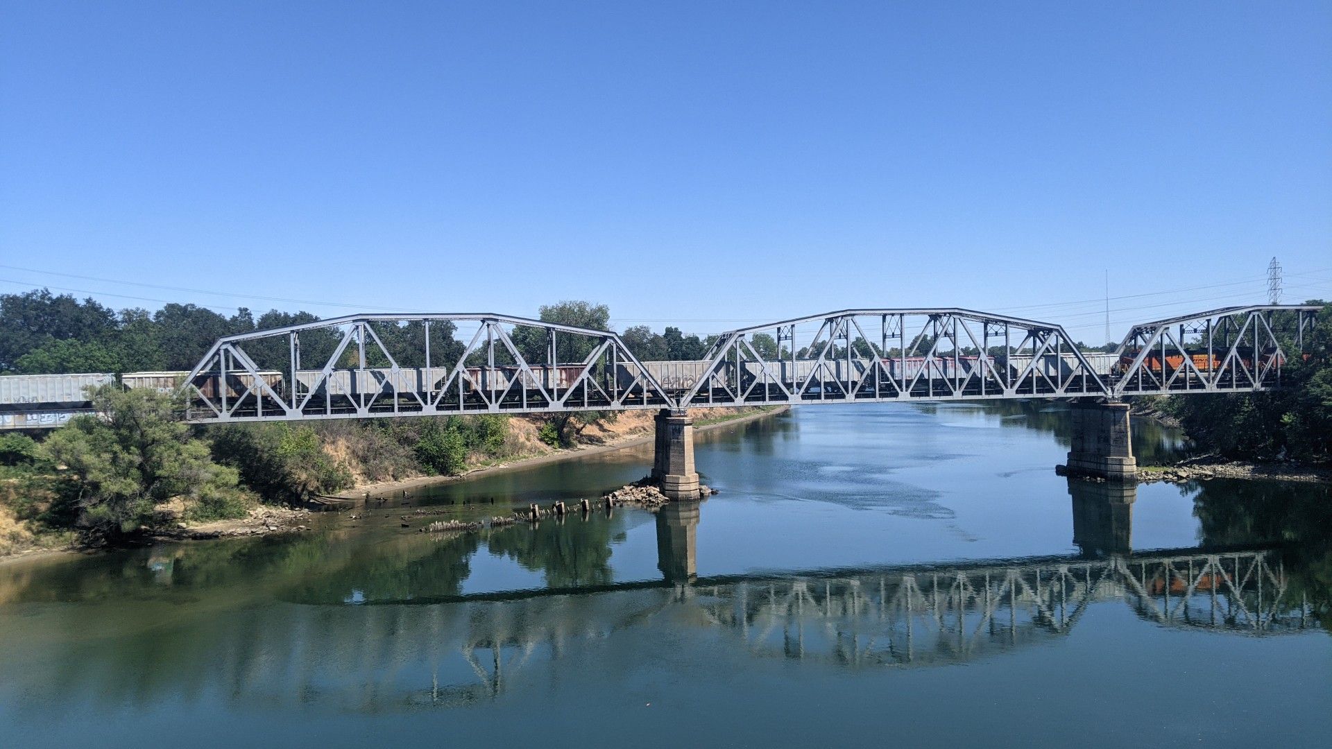 A long metal truss bridge crosses a calm river under a bright blue sky. - American River and Sacramento River Trail Loop