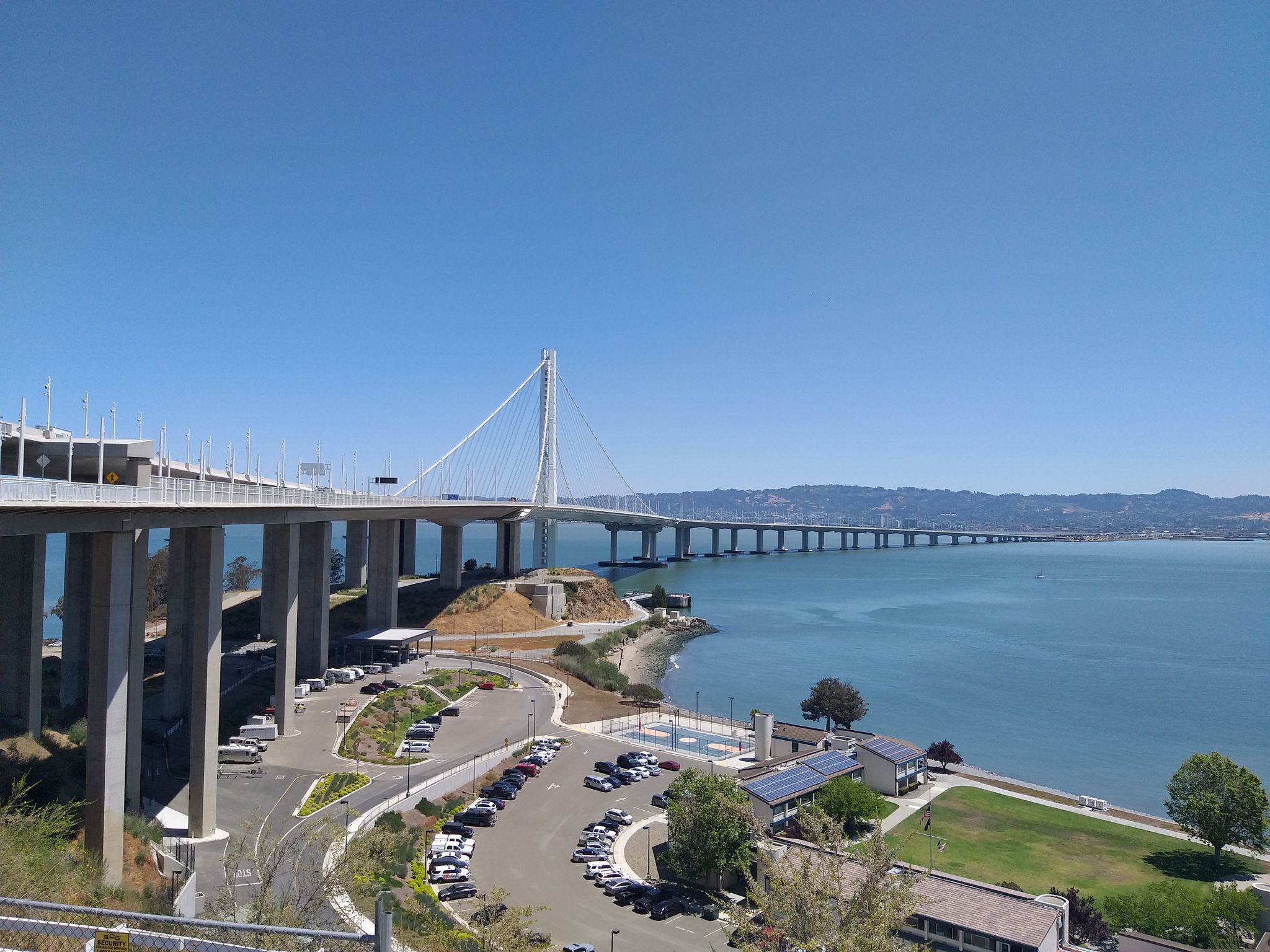 Large suspension bridge over water, with a parking lot and buildings below. Bright day. - Bridge Yard – Emeryville Bayfront Bridge loop from Emeryville