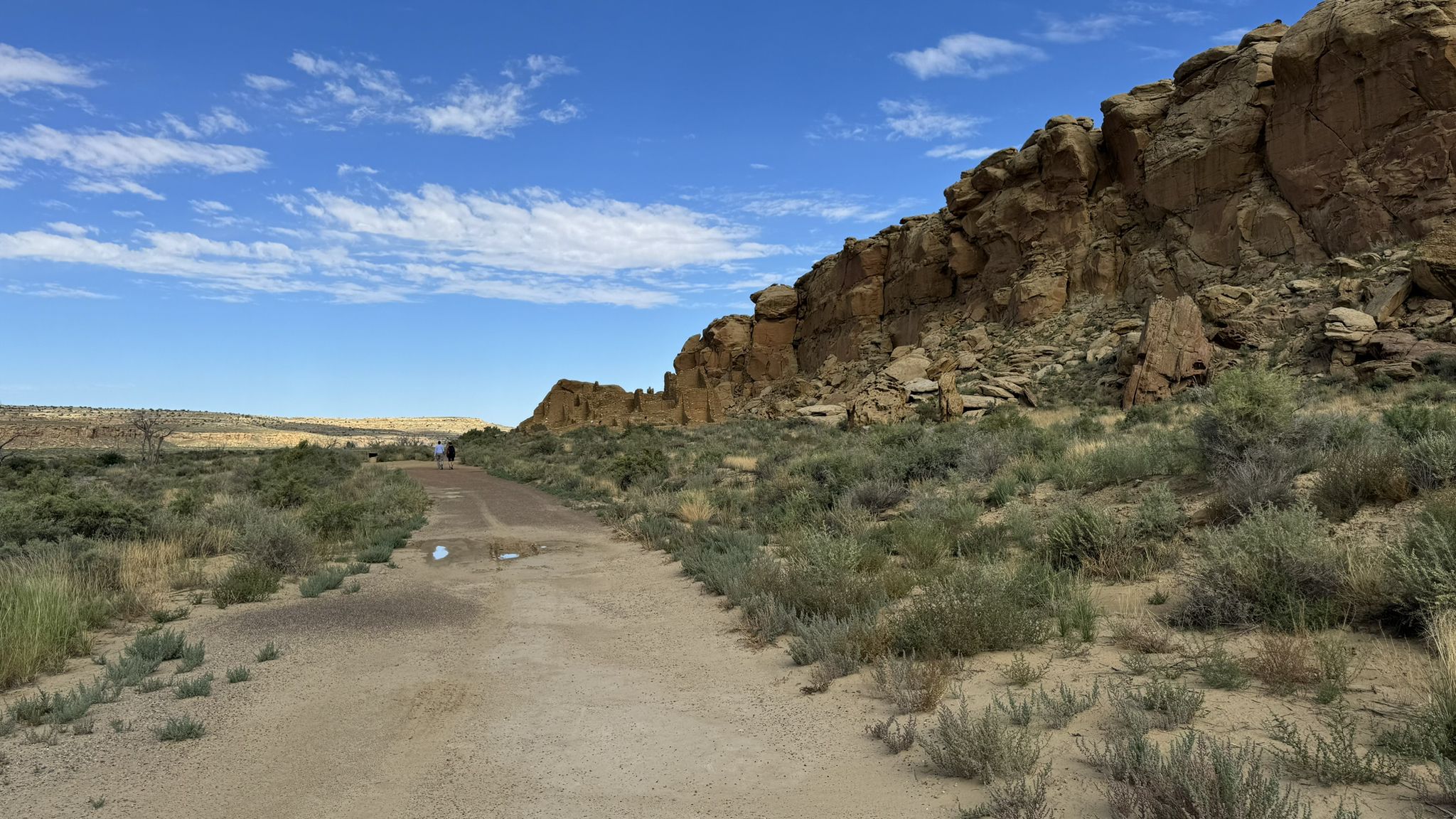 Dirt path alongside a rocky cliff in a desert landscape under blue sky. - Kin Kletso Trail