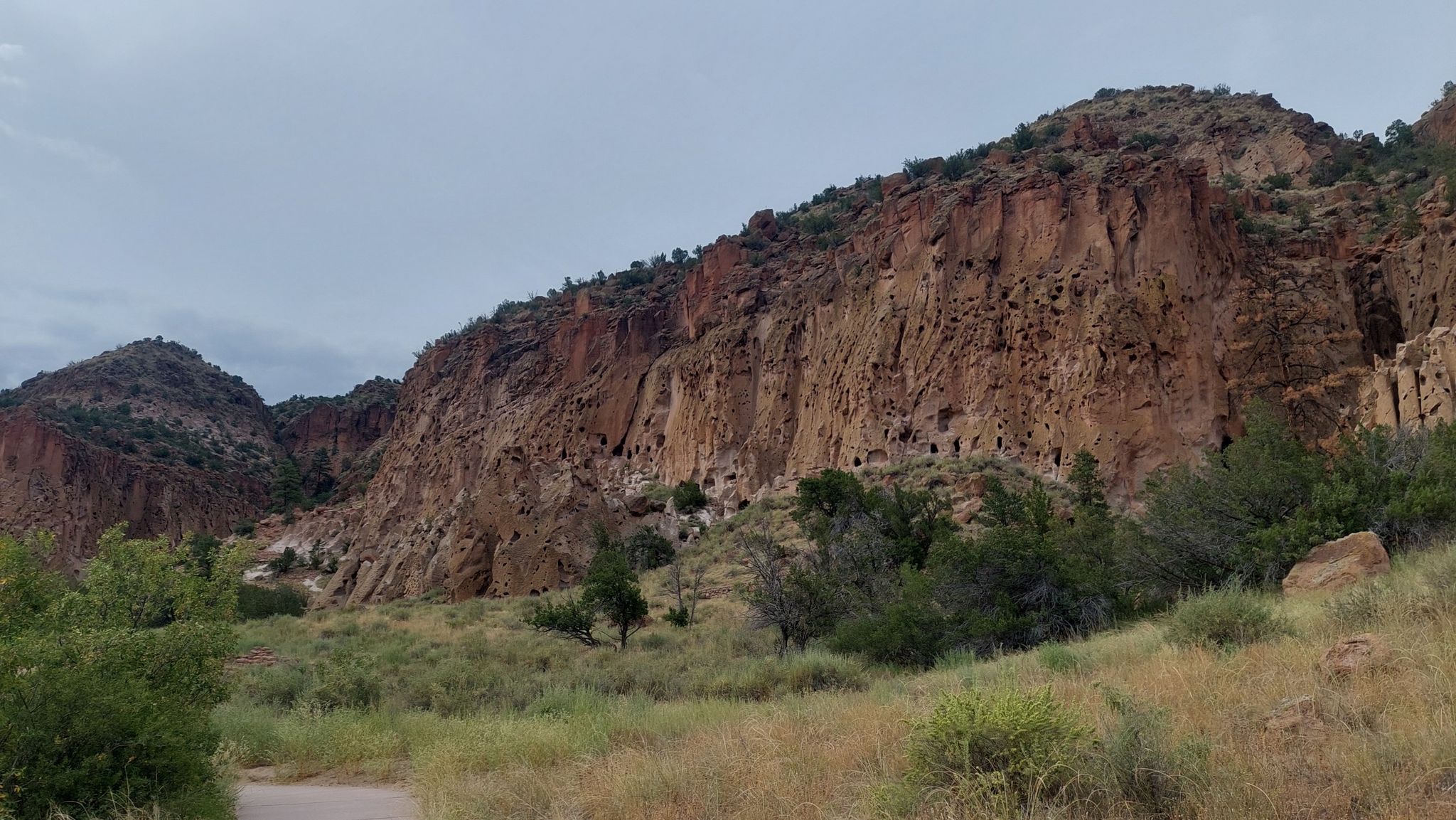 Massive rocky cliff face with many small caves under an overcast sky. - Pueblo Loop Trail