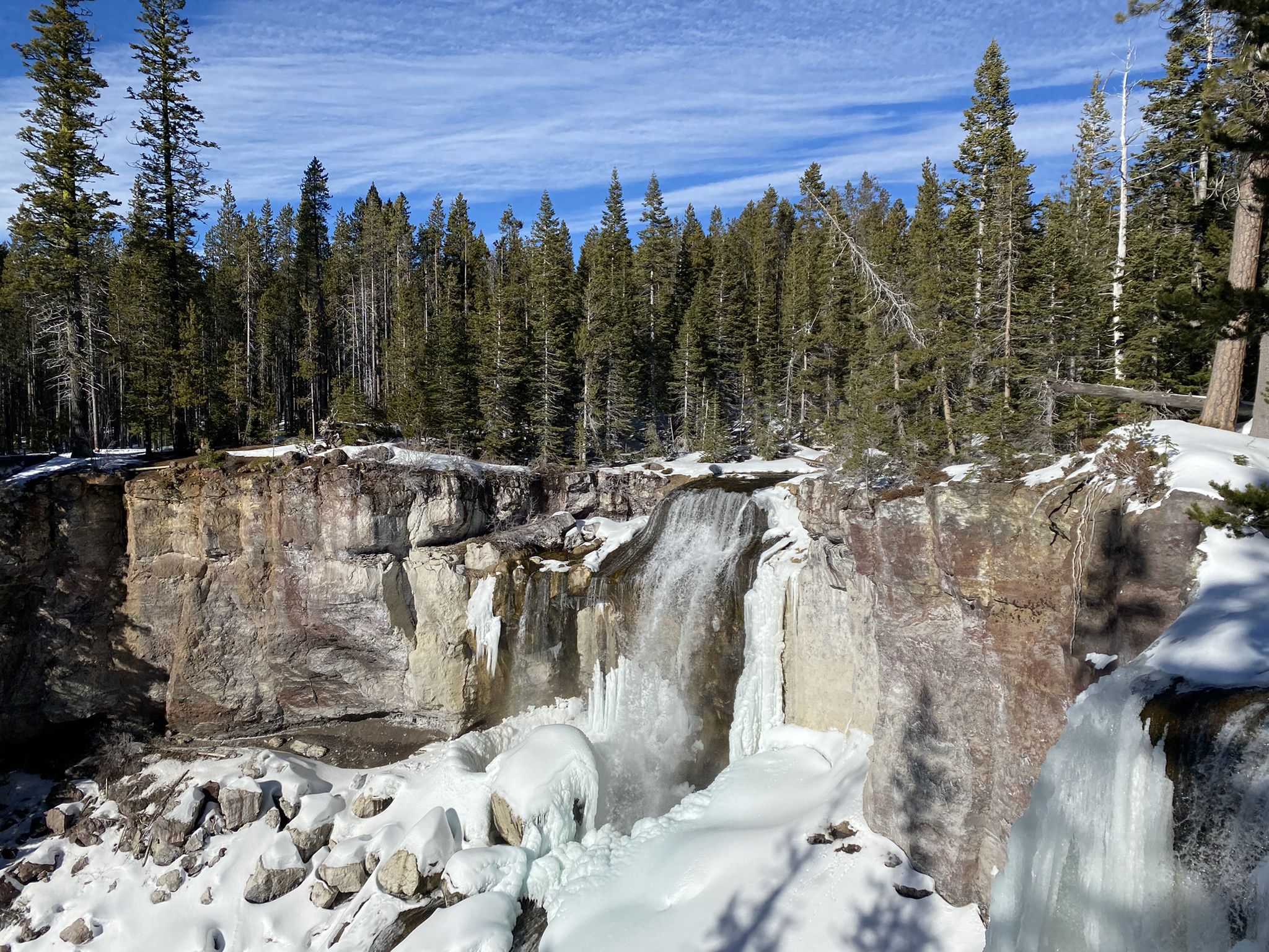 A powerful waterfall cascades over icy rocks into a snowy canyon with pine trees. - Paulina Creek Falls Trail