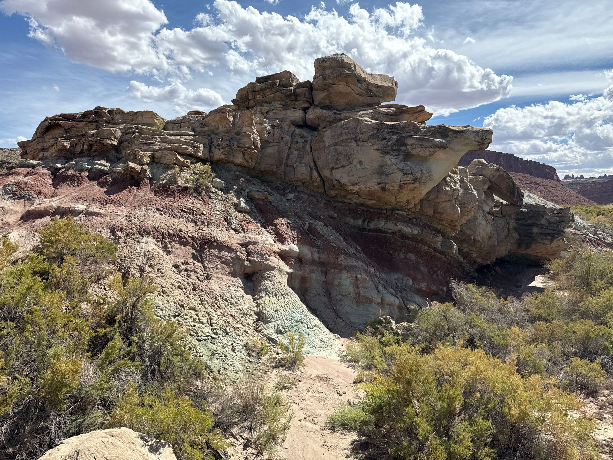 Colorful layered rock formation with green shrubs under a partly cloudy sky. - Delicate Arch Trail